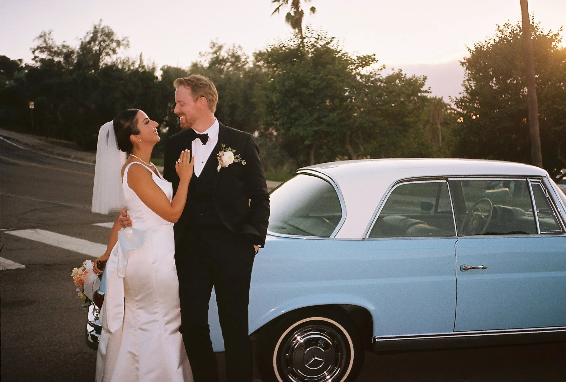 A bride and groom in wedding attire smiling and looking at each other beside a vintage blue car during sunset.