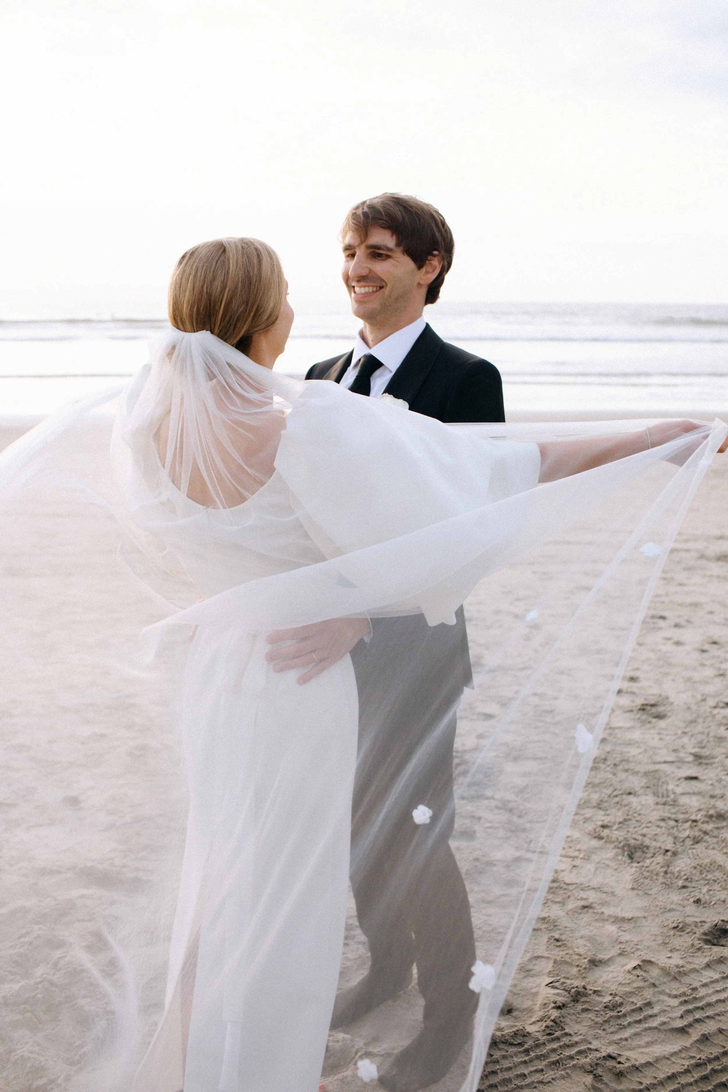 A newly married couple on the beach, with the groom smiling at the bride who is wrapped in a sheer veil, during sunset or early evening.
