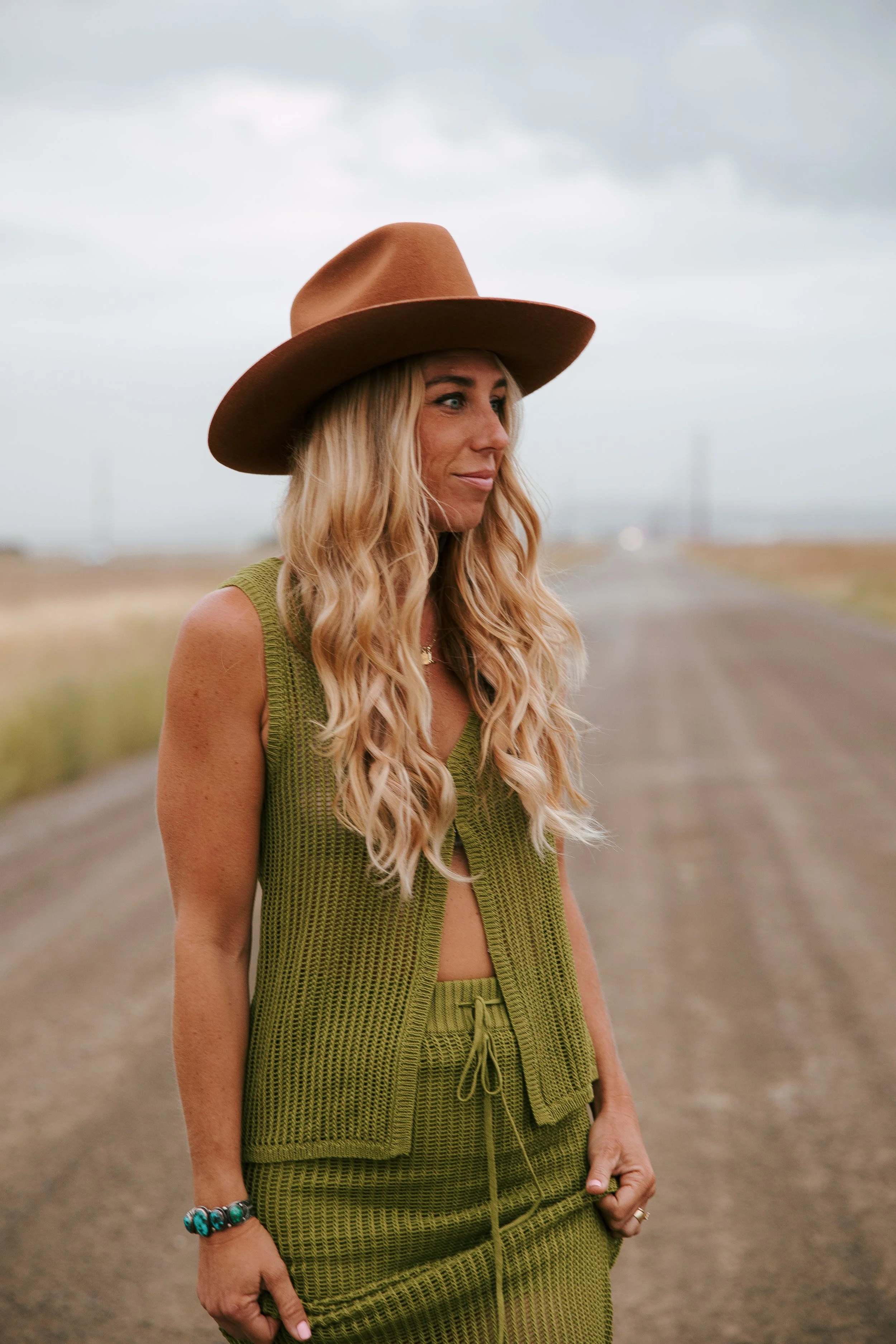 A woman with long blonde curly hair wearing a wide-brimmed brown hat, green sleeveless top, and matching green shorts, standing on a rural dirt road under a cloudy sky, with her right arm relaxed and her left hand holding the waistband of her shorts.