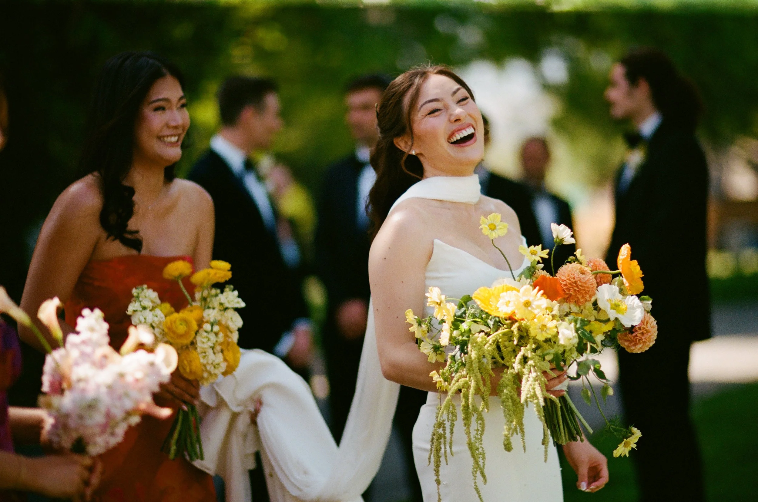A group of people at a wedding, with women holding bouquets of flowers, smiling and laughing outdoors under green trees.