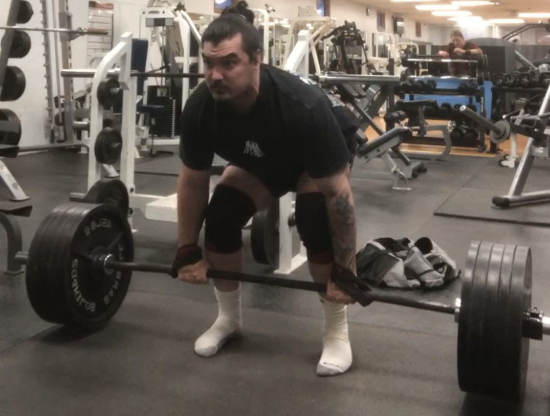 A man in a gym performing a deadlift exercise with a loaded barbell, wearing knee sleeves and gloves, with gym equipment and weights in the background.
