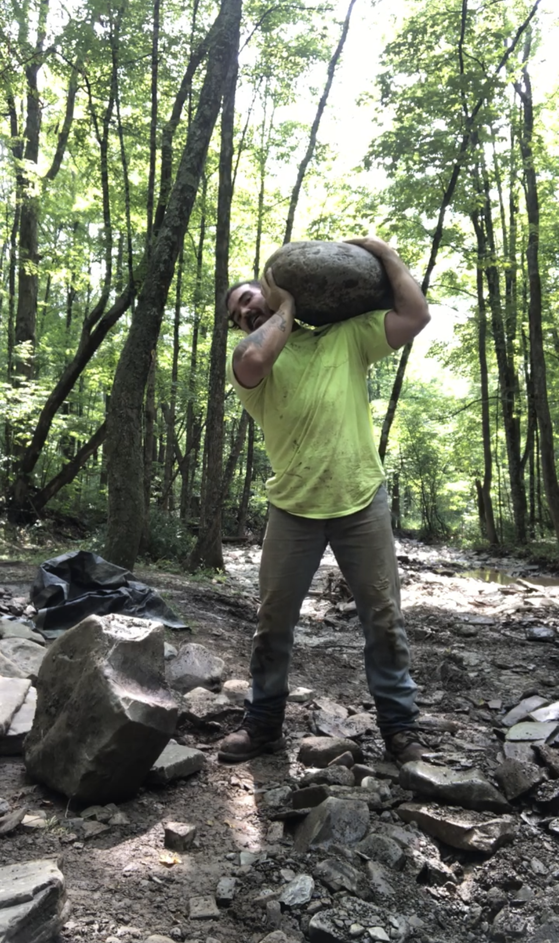 A man wearing a yellow shirt and jeans is lifting a large rock in a forested area, with trees and a stream in the background.