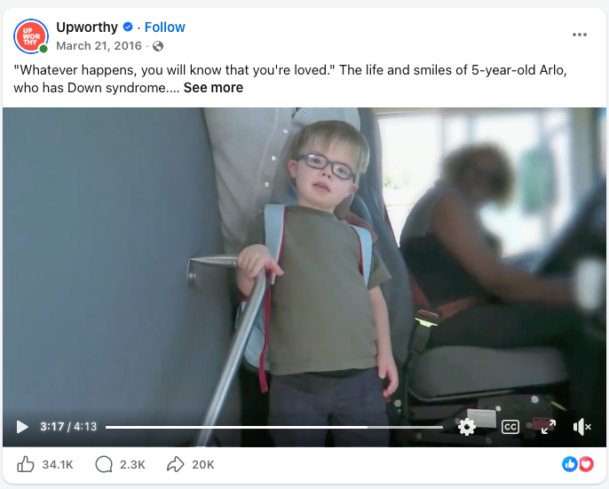 A young boy with Down syndrome stands on an airplane with the assistance of a rail, smiling softly, with a blurred person sitting nearby.