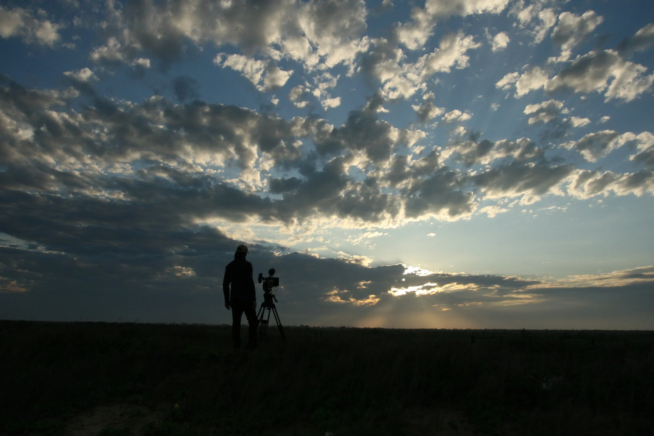 Person standing next to a camera on a tripod outdoors during sunset or sunrise with a partly cloudy sky.