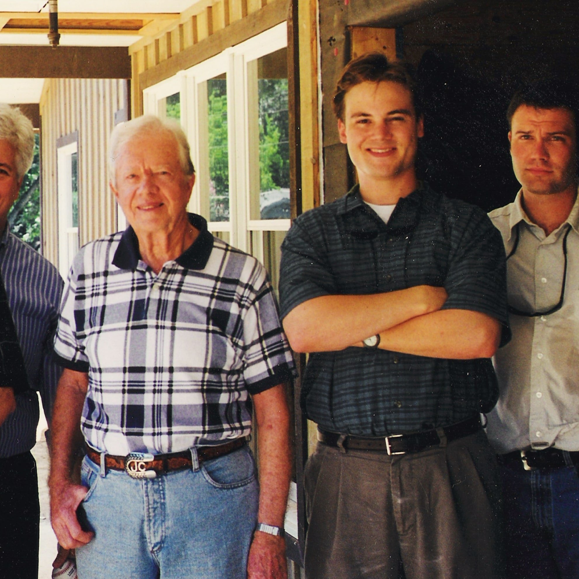 Four men standing side by side, smiling at the camera, outdoors near a partially constructed building with windows and wooden framing.