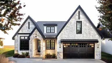 Front view of a modern house with a gabled roof, white brick exterior, black window frames, and a black garage door, surrounded by trees.