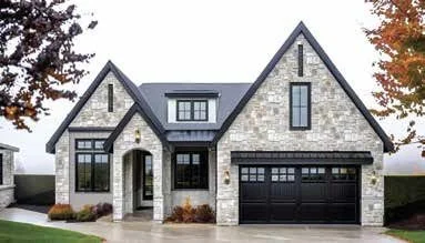 Front view of a modern stone house with a garage, large windows, and a peaked roof, autumn trees in the background.
