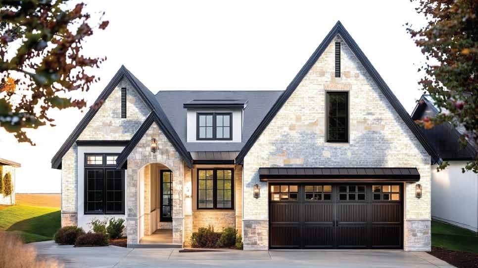 A modern two-story house with a stone and stucco exterior, black garage door, and multiple pitched roofs, surrounded by a well-maintained lawn and trees.