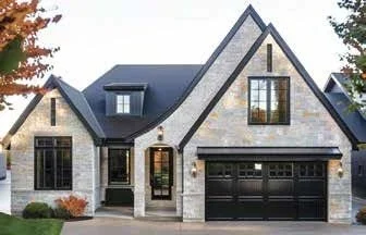 Exterior view of a modern house with a dark roof, stone and brick facade, large windows, and a black garage door, surrounded by trees.