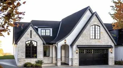 Front view of a modern two-story house with a stone facade, black roof, arched windows, and attached garage, surrounded by trees with autumn leaves.