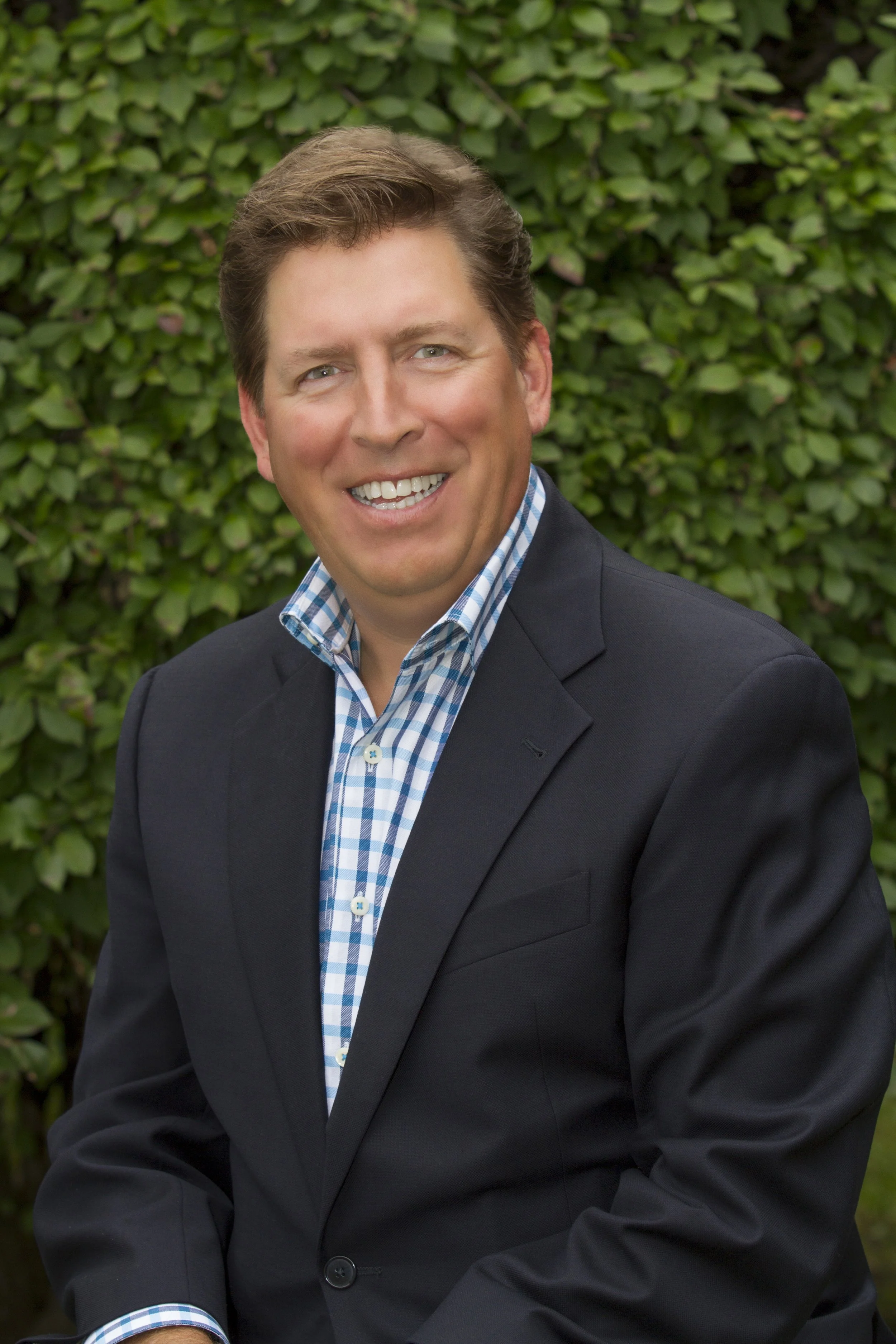 A man in a dark suit with a checkered shirt, smiling, outdoors with greenery background.