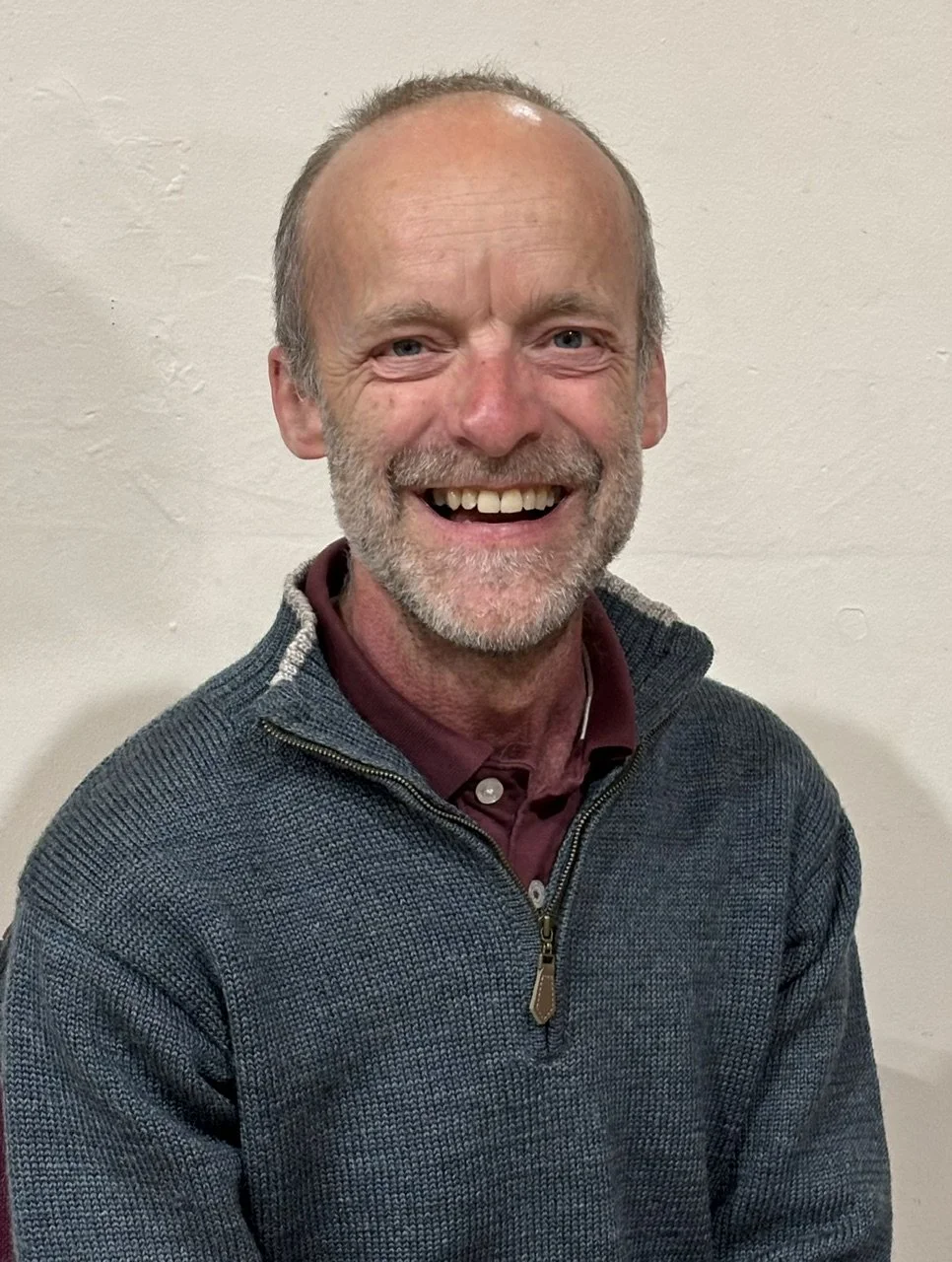 A smiling middle-aged man with a beard and short hair, wearing a gray zip-up sweater over a maroon collared shirt, standing against a plain light-colored wall.