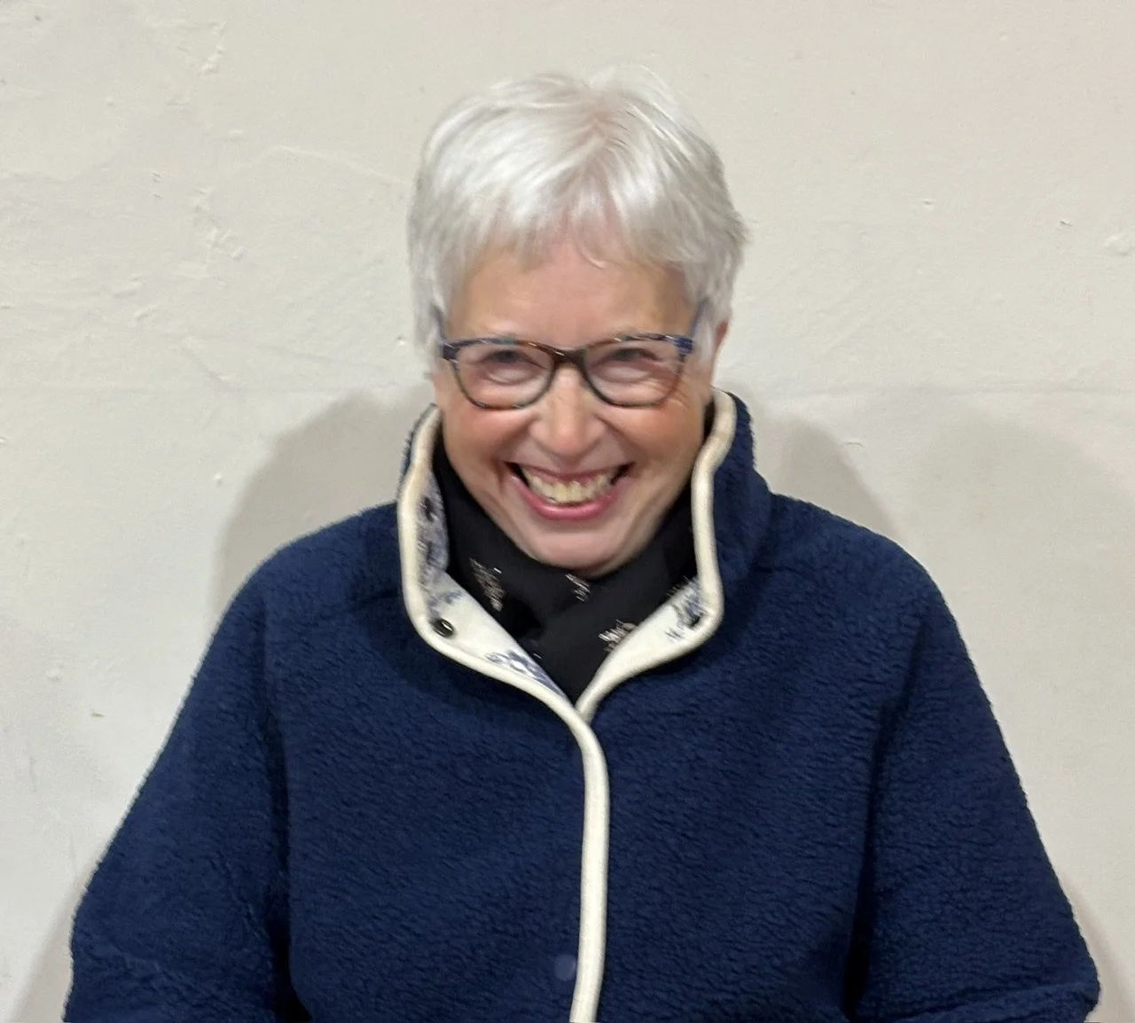A smiling elderly woman with short gray hair and glasses, wearing a navy blue fleece jacket and a black shirt, standing against a plain white wall.