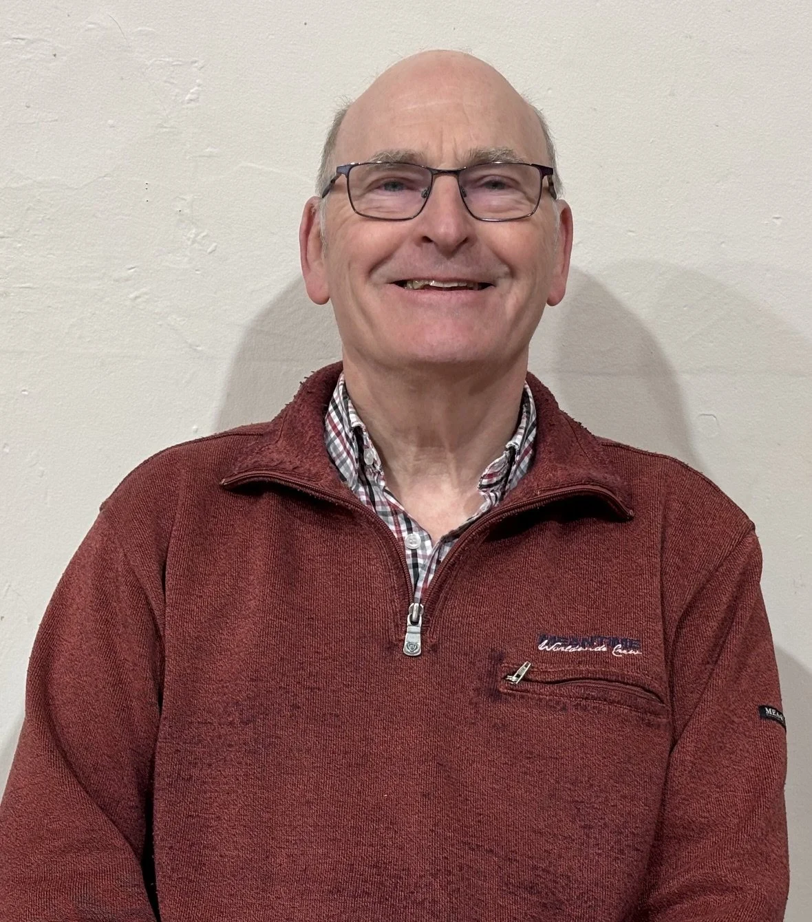 A smiling elderly man wearing glasses, a red quarter-zip sweater, and a plaid shirt standing against a plain wall.
