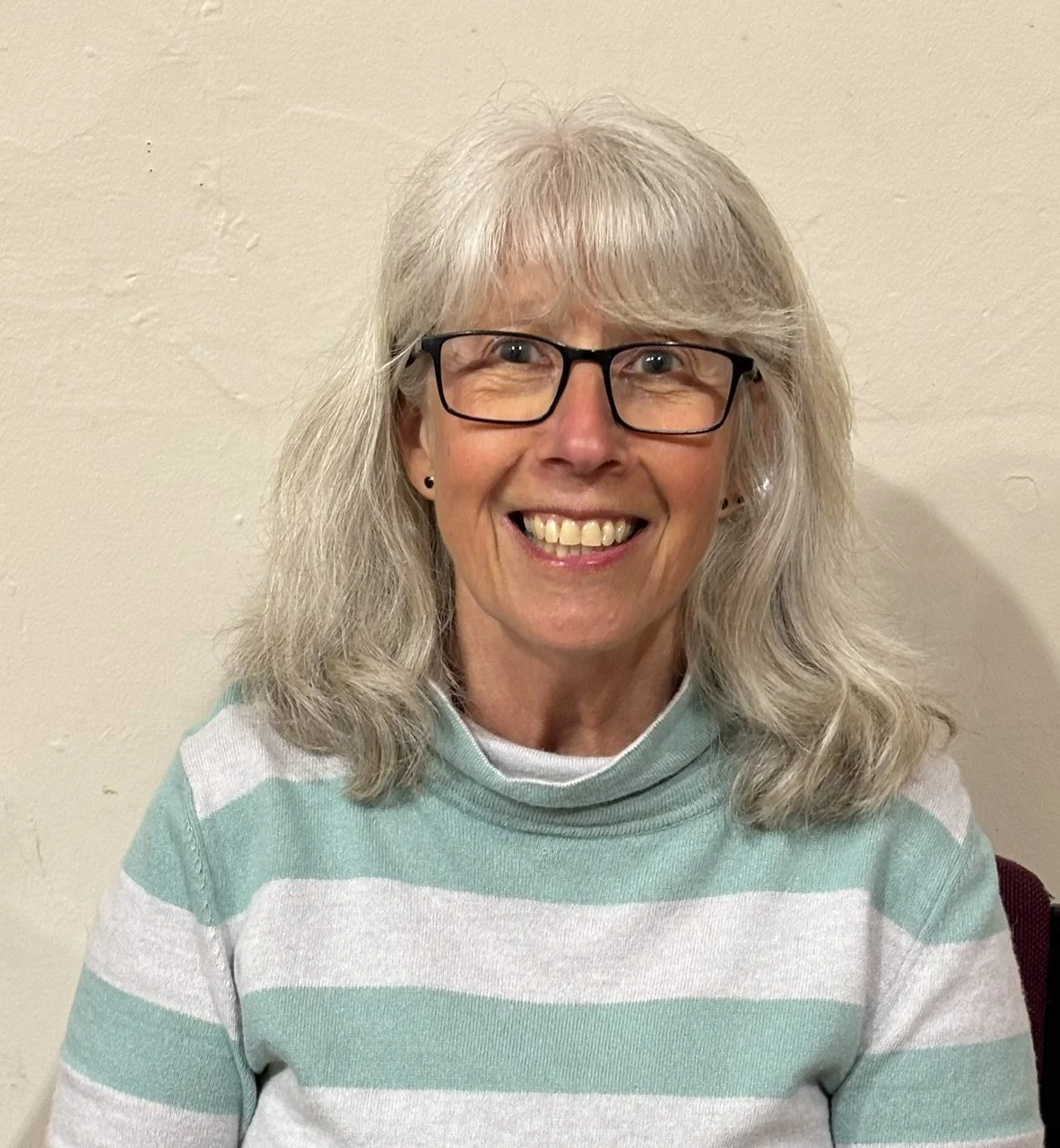 A smiling woman with gray hair, wearing glasses and a mint green and white striped sweater, standing against a plain wall.