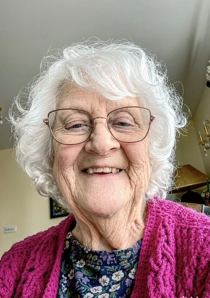 Portrait of an elderly woman with white curly hair, wearing glasses, a floral blouse, and a magenta sweater, smiling cheerfully indoors.