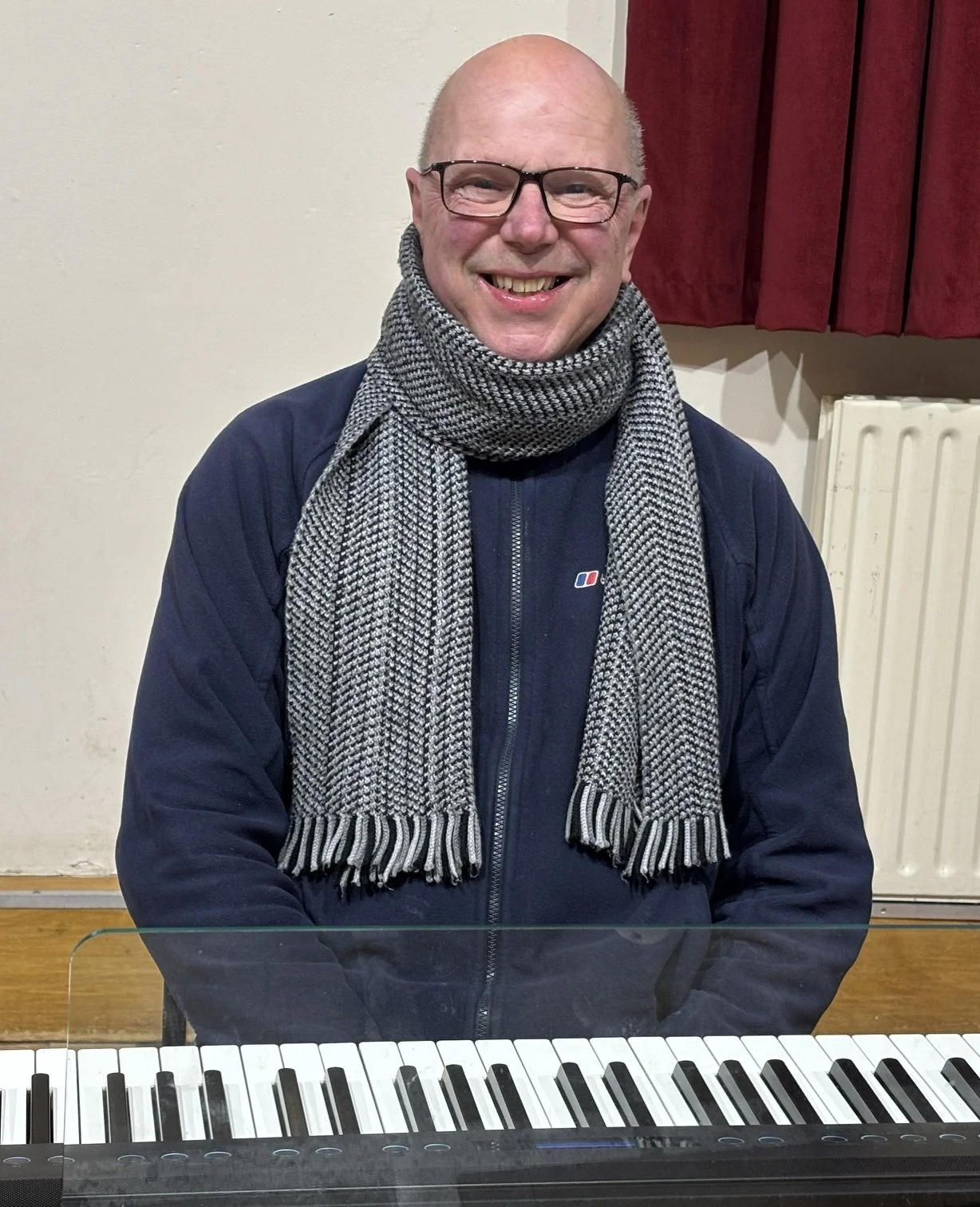 A smiling man with glasses sitting behind a keyboard. He is wearing a dark jacket and a gray and white striped scarf. The background includes a red curtain and a white radiator.