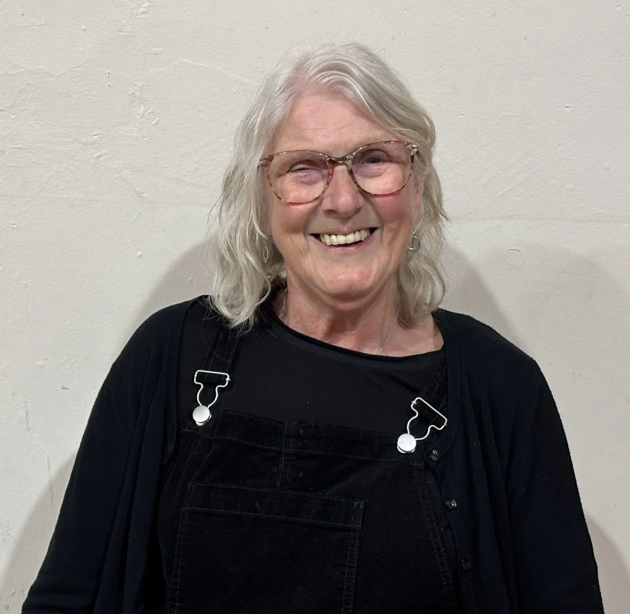 A smiling elderly woman with glasses, wavy gray hair, wearing a black shirt and overalls, standing against a plain white wall.