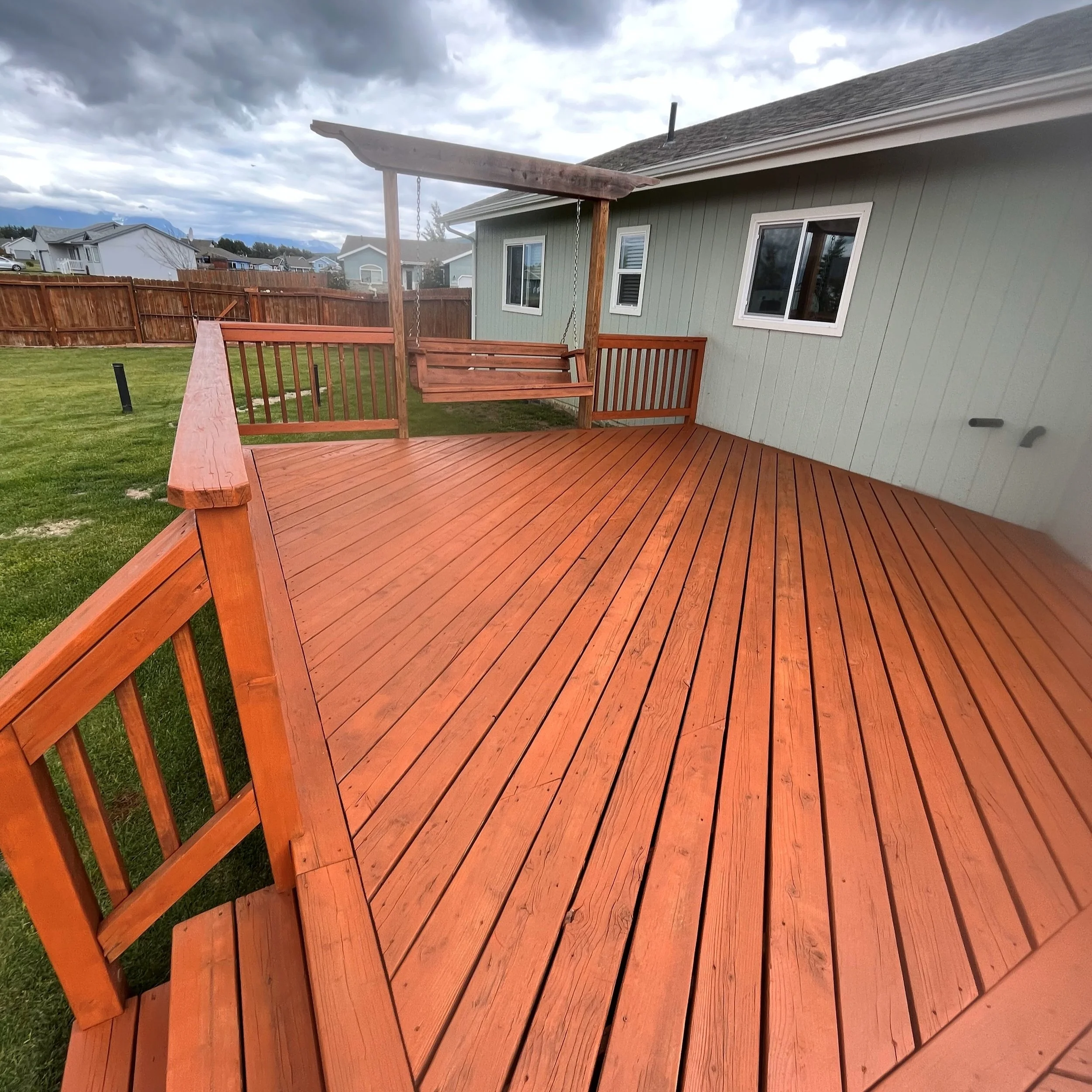 A wooden deck attached to a house, with a swing seat under a partial roof, overlooking a grassy backyard enclosed by a wooden fence, with suburban houses in the background and cloudy skies above.