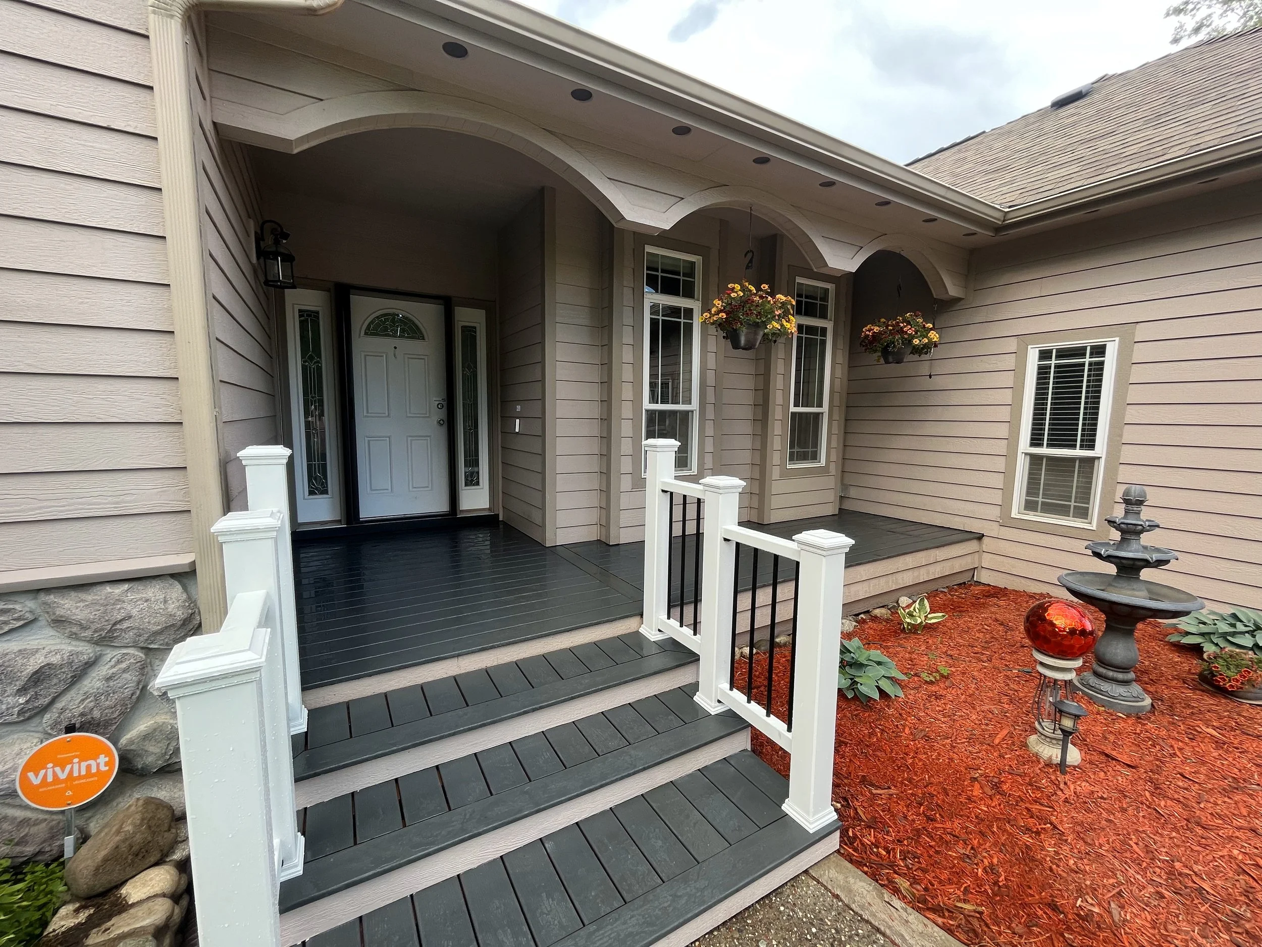 Front porch of a house with dark gray stairs, white railings, hanging flower baskets, and a black fountain surrounded by mulch and plants