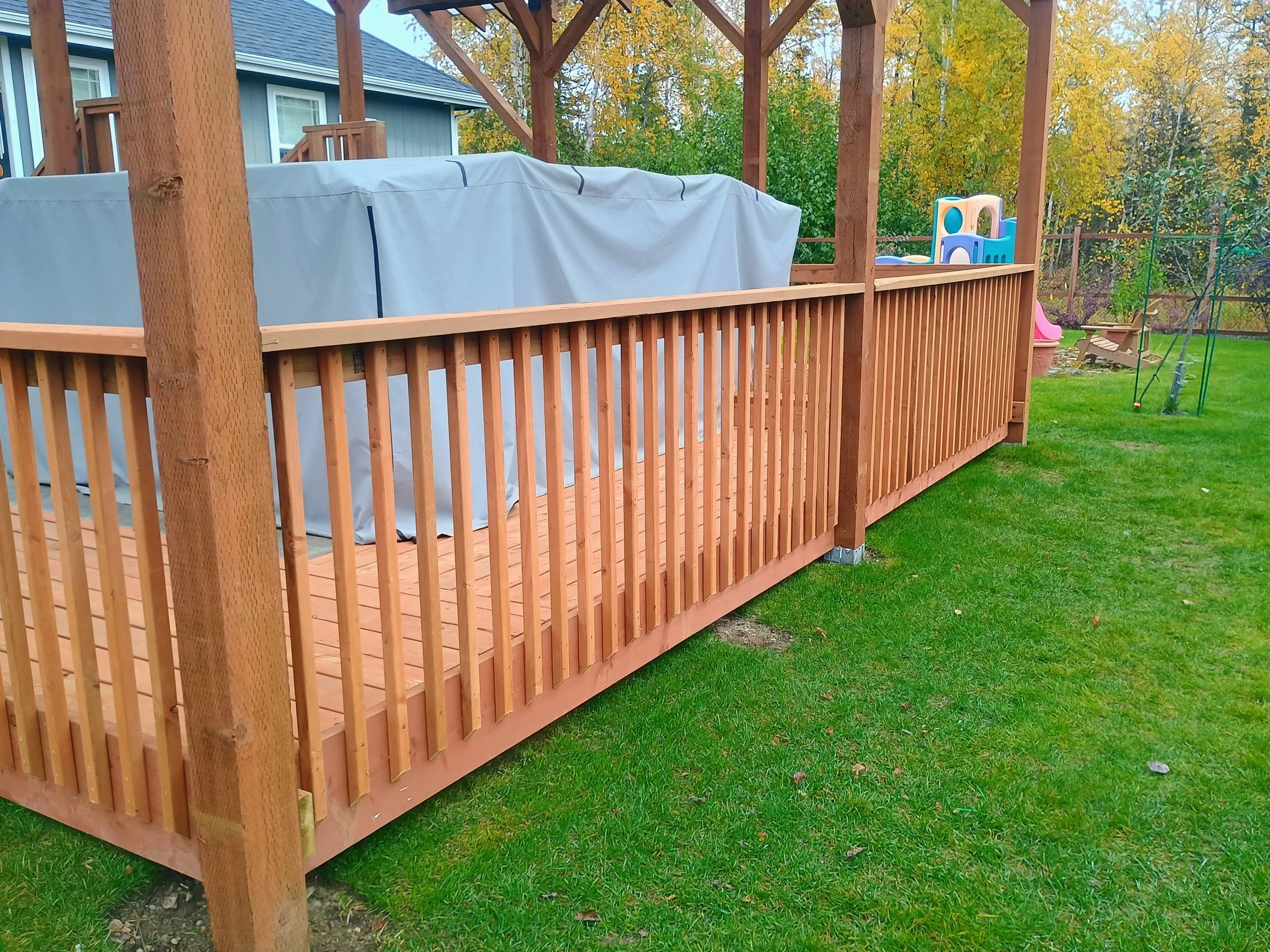 A wooden backyard deck with protective railing, several vertical posts, and a covered area, surrounded by a green lawn and trees with autumn foliage, with a blue house visible in the background.