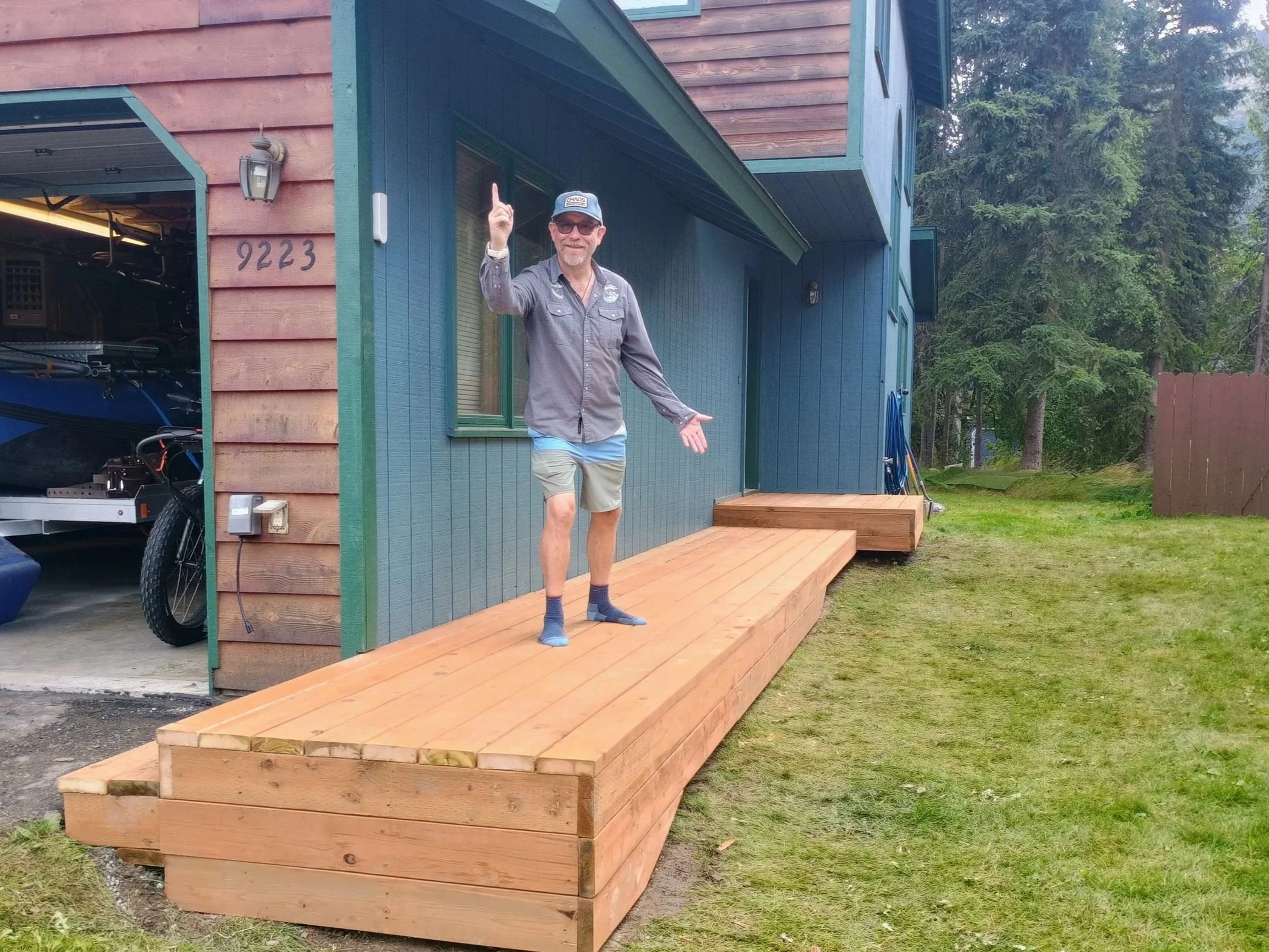 A man is standing on a newly built wooden deck outside a blue house with green trim. The man is smiling, wearing sunglasses, a gray cap, a gray shirt, shorts, and socks. Some trees and grass are visible in the background.