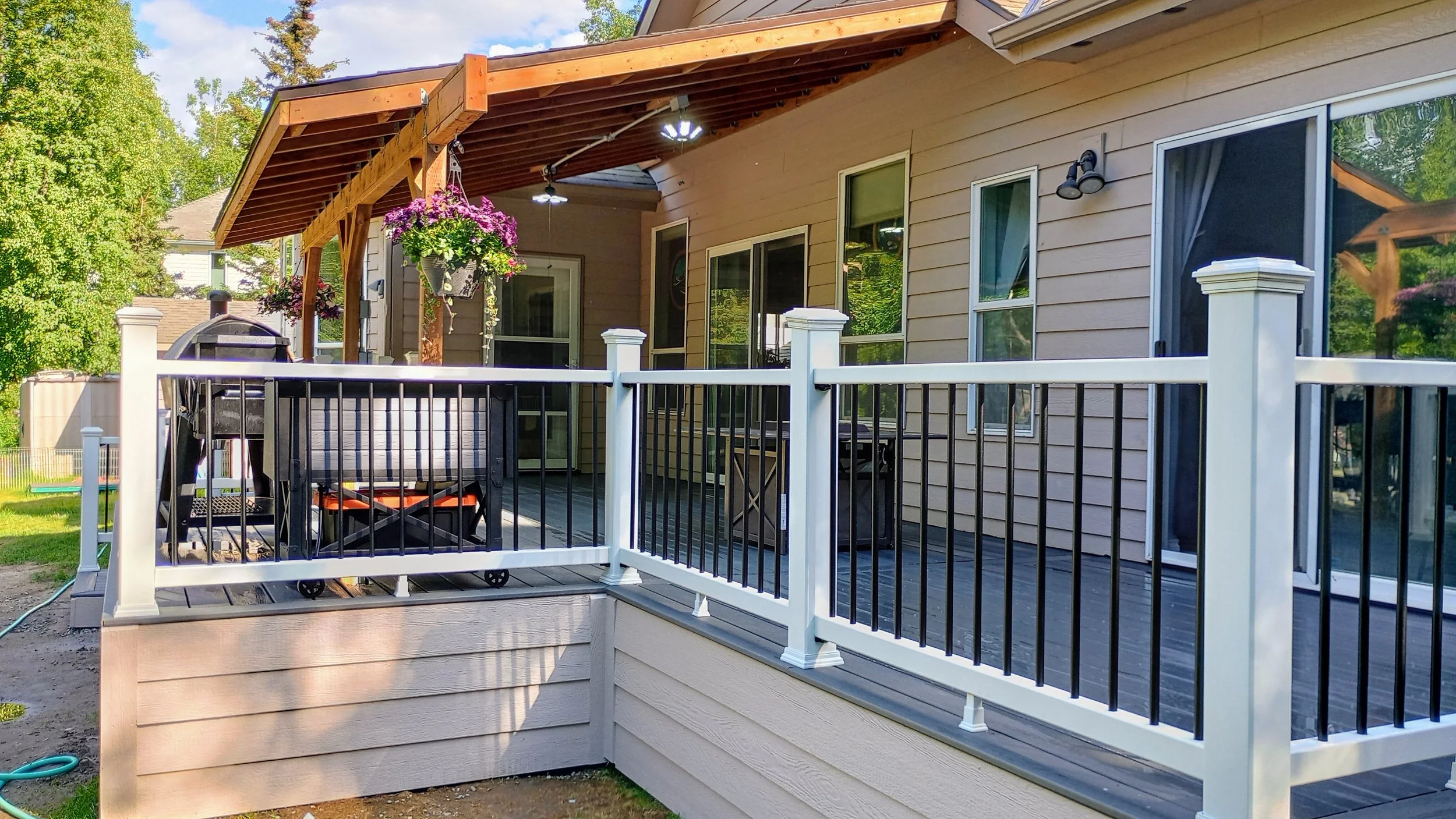 Backyard deck with black and white railing, hanging flower baskets, outdoor furniture, grill, and a house with beige siding and multiple windows, surrounded by green trees.