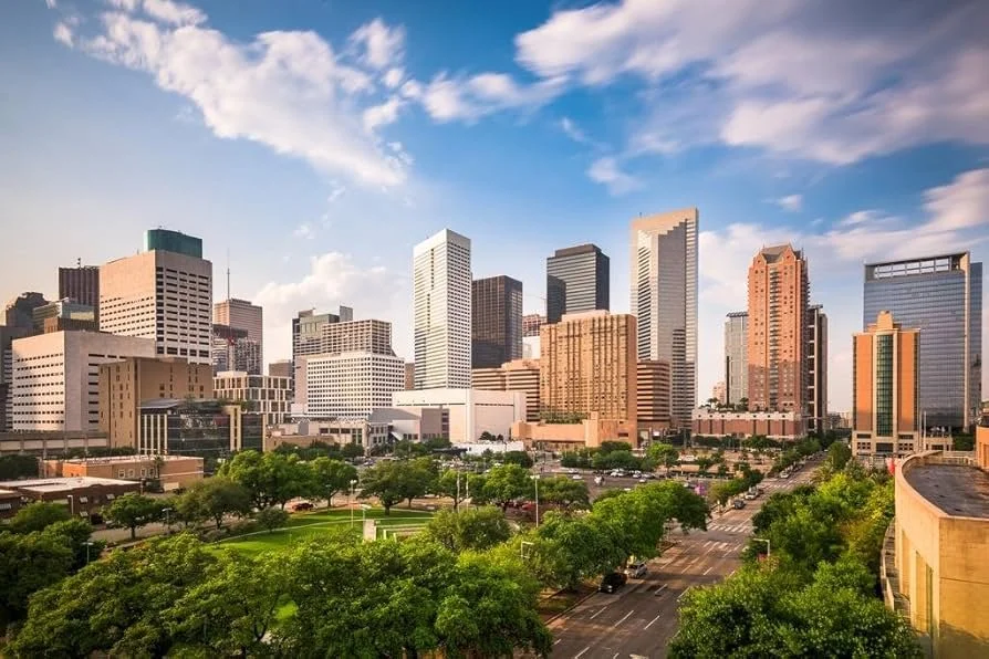 A high-angle view of a sunlit city skyline with modern skyscrapers. A lush green park with dense trees sits in the foreground under a bright blue sky with wispy white clouds.