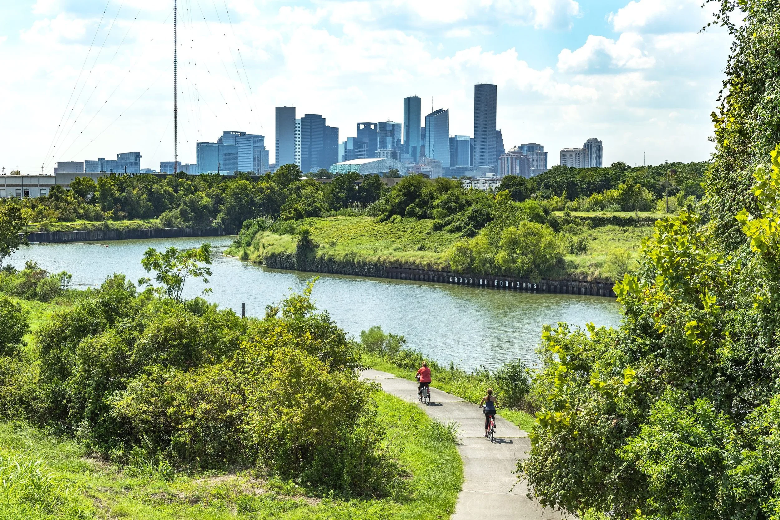 Two people bike along a paved river path. Vibrant green shrubbery lines the water, while a distant city skyline is visible against a bright, hazy sky, blending nature and urban life.