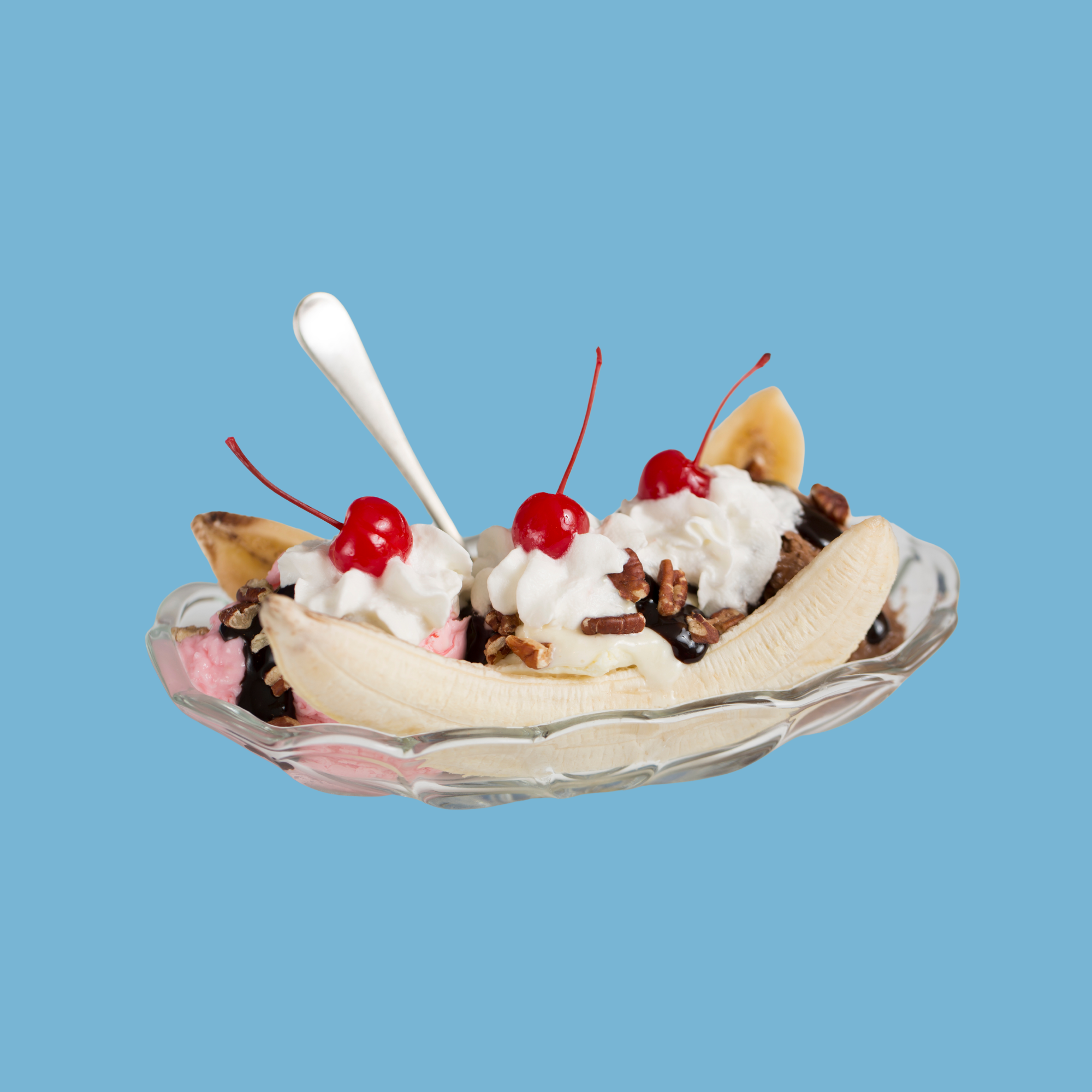 A glass dish with sliced bananas, strawberries, whipped cream, chocolate syrup, and nuts, topped with cherries and a spoon, against a light blue background.