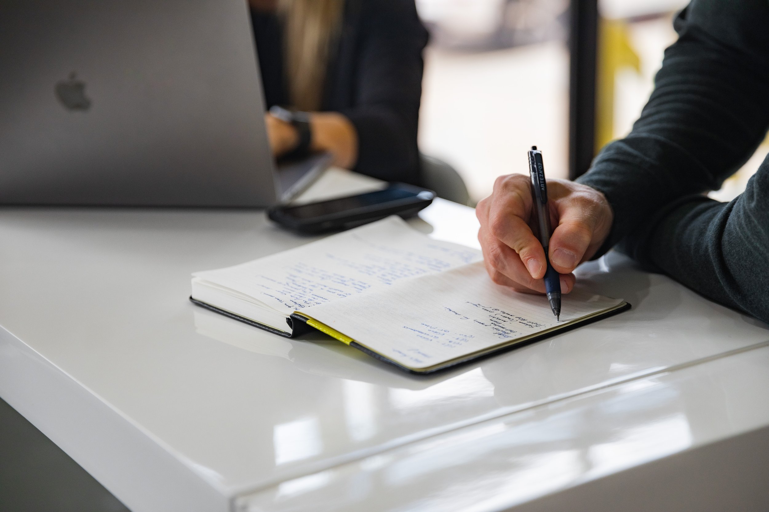 Person writing notes in a notebook at a desk with a laptop and smartphone.