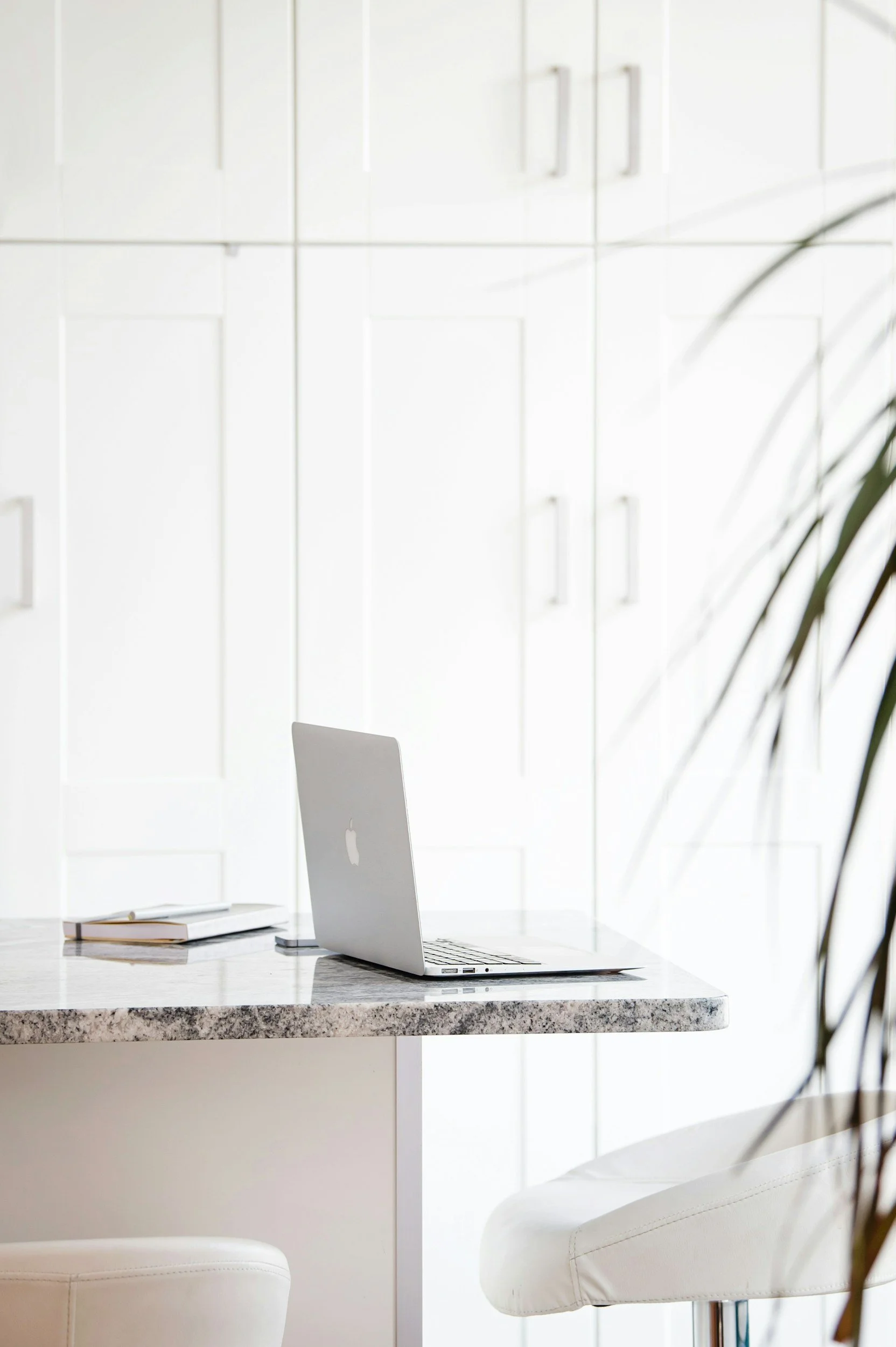A minimalist home office with a marble top table, a silver MacBook, a closed notebook, and a white chair, with white cabinetry and a potted plant partially visible.