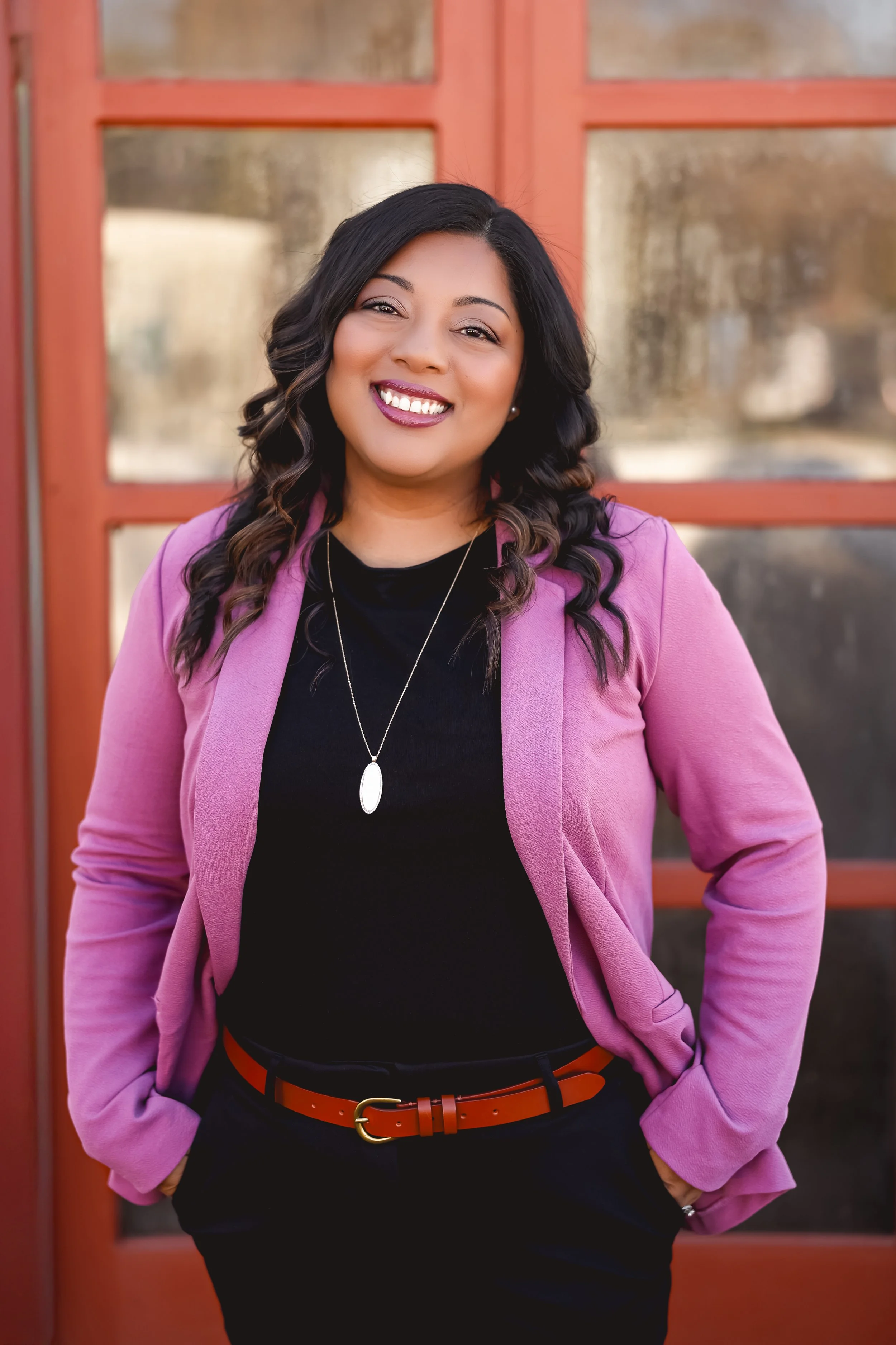 A woman with shoulder-length dark wavy hair, smiling, wearing a purple blazer over a black top, with a red belt, standing in front of a red-framed window.