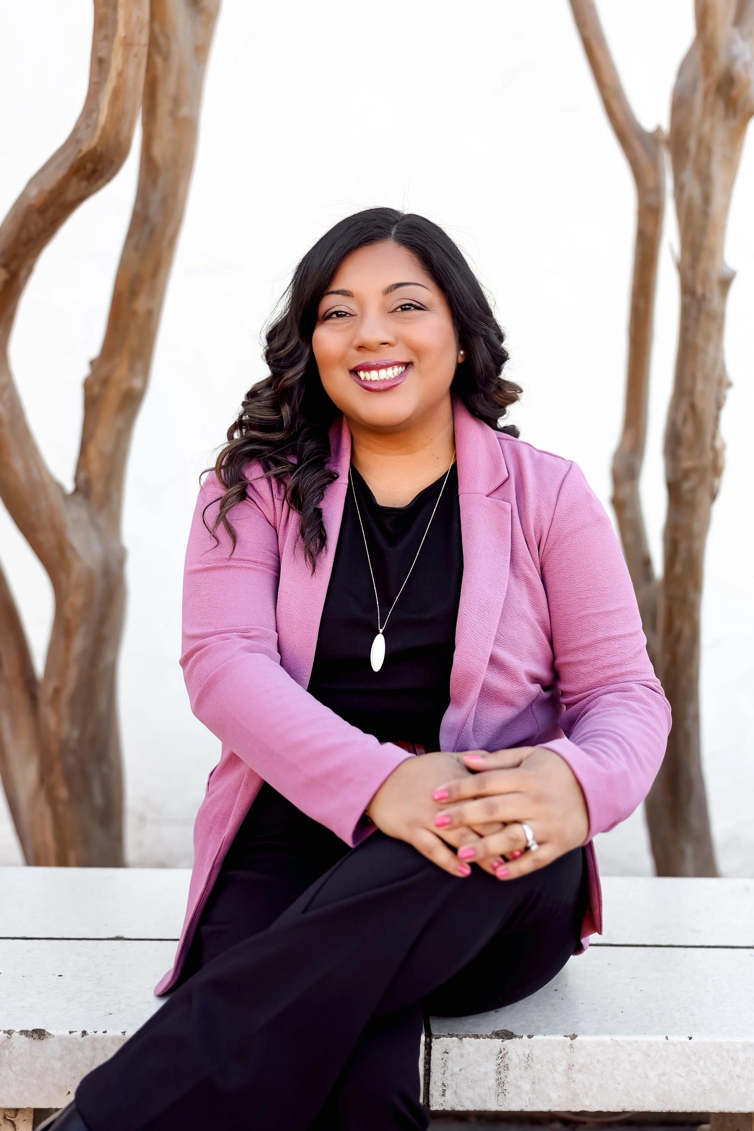 A woman sitting on a white bench outdoors, wearing a pink blazer over a black top, with a necklace, smiling, with trees in the background.
