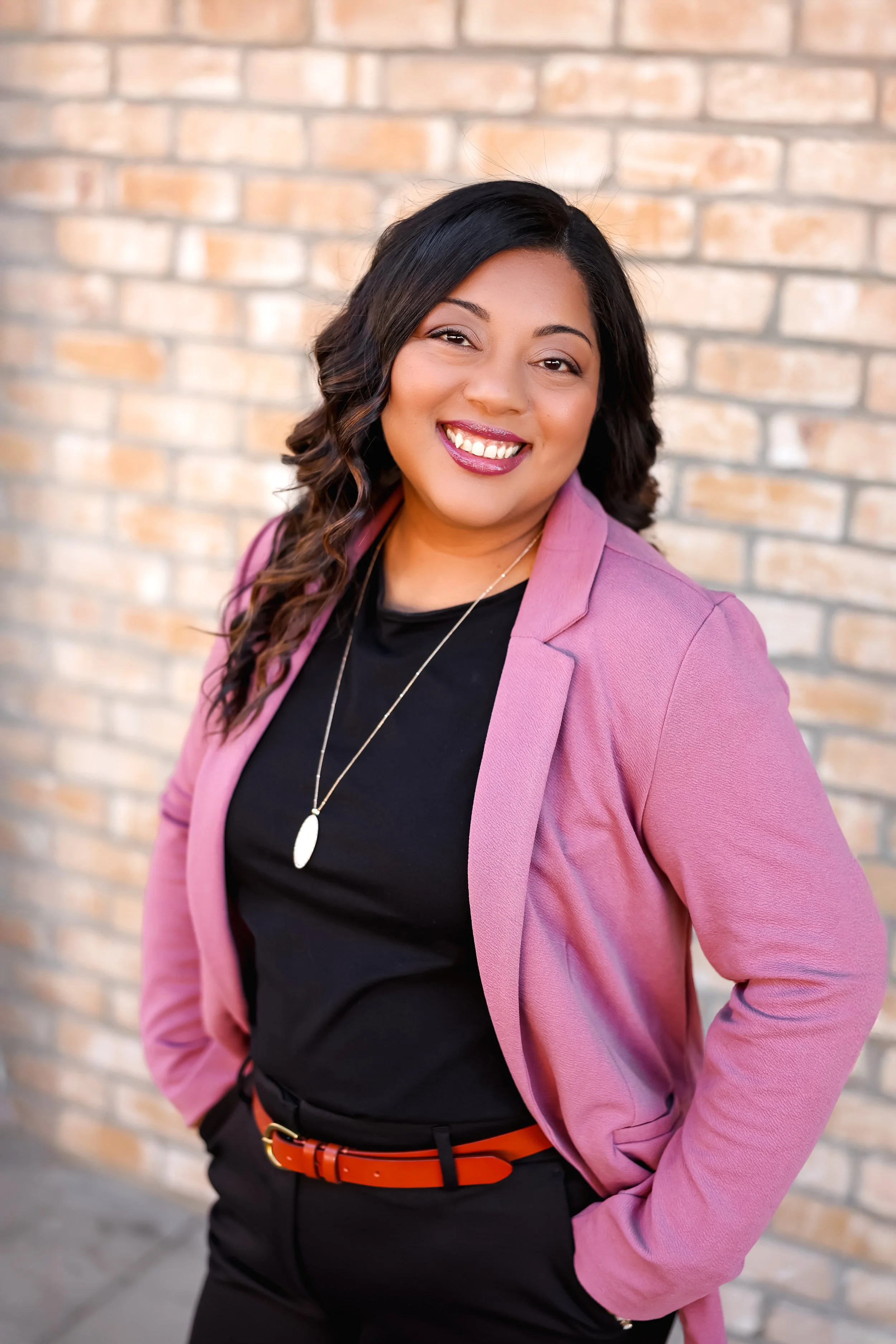 A woman with long dark hair wearing a pink blazer, black top, orange belt, and a necklace, smiling against a brick wall background.