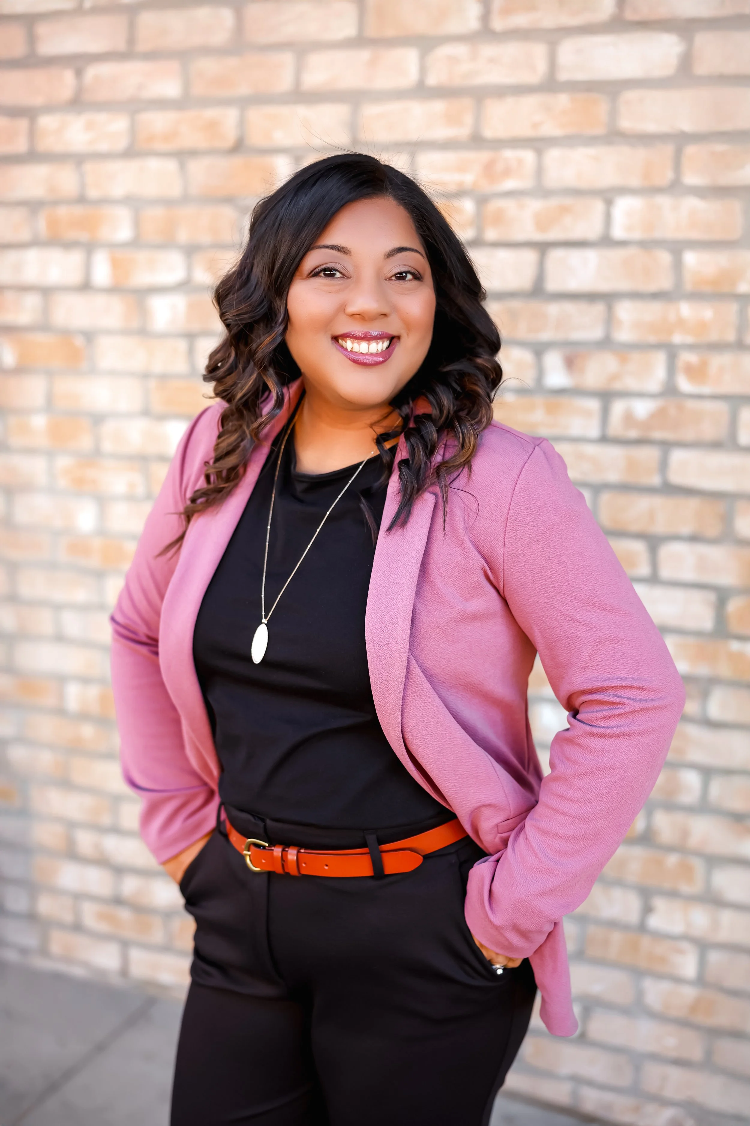 Portrait of a smiling woman with dark hair wearing a pink blazer, black shirt, and black pants with an orange belt, standing against a brick wall.