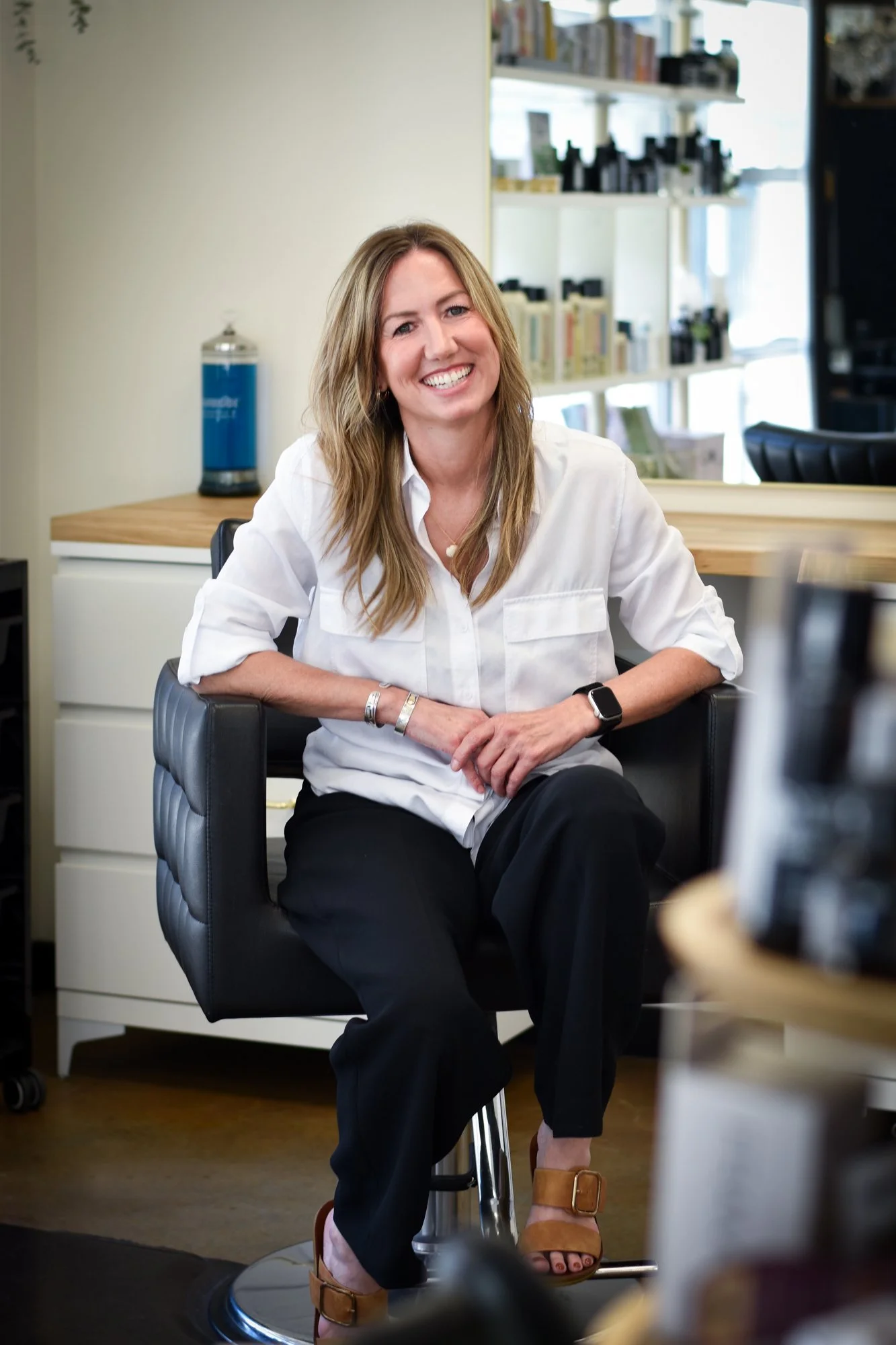 A woman with long blonde hair, smiling, sitting on a black chair in an office or studio. She is wearing a white blouse, black pants, tan heels, and a smartwatch. There's a white drawer cabinet and a water dispenser behind her, with shelves and shelves of bottles and books in the background.