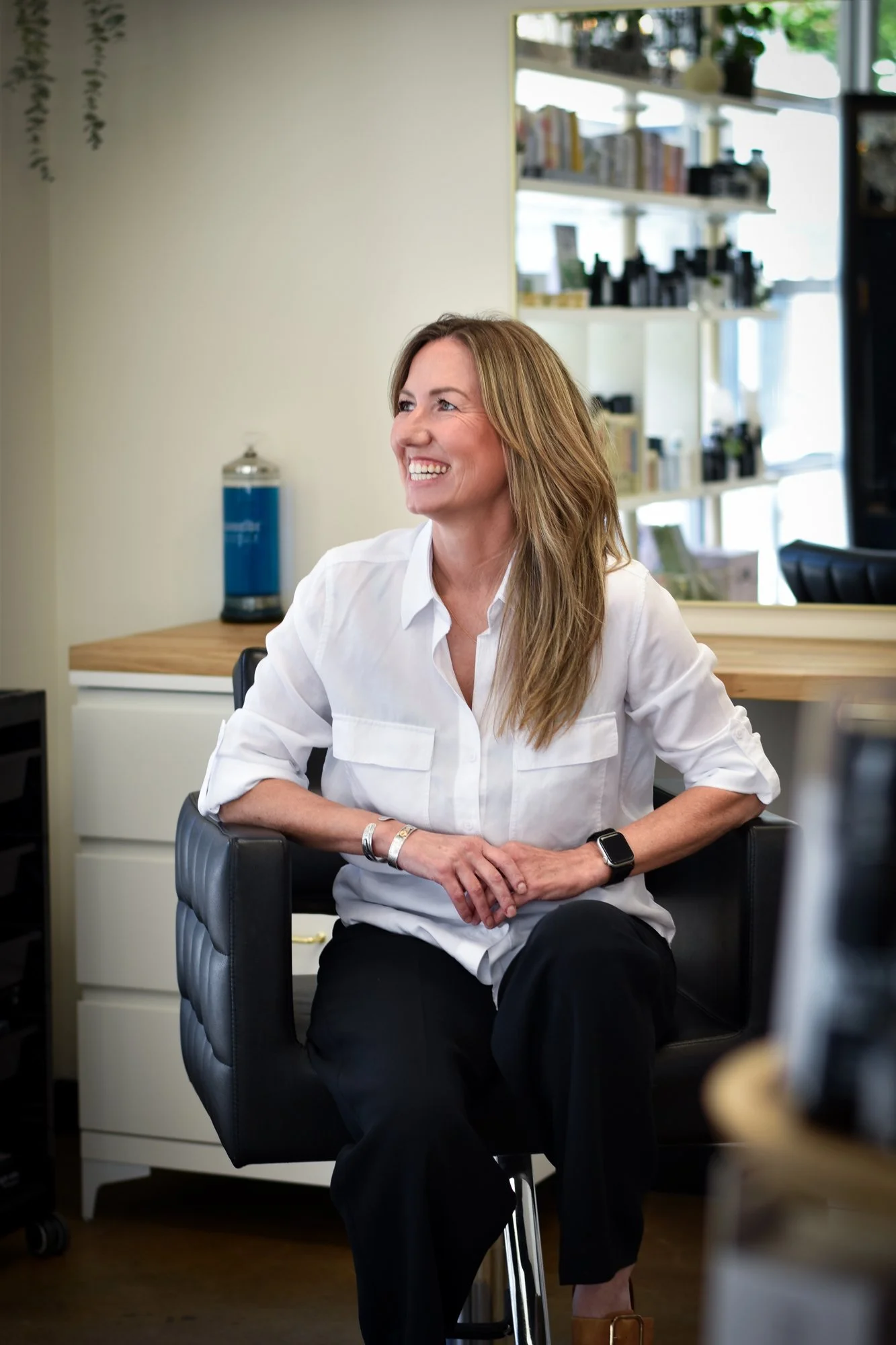 A smiling woman with long wavy hair, wearing a white button-down shirt and black pants, sitting on a black office chair in a modern office space with a bookshelf, a water dispenser, and large windows in the background.