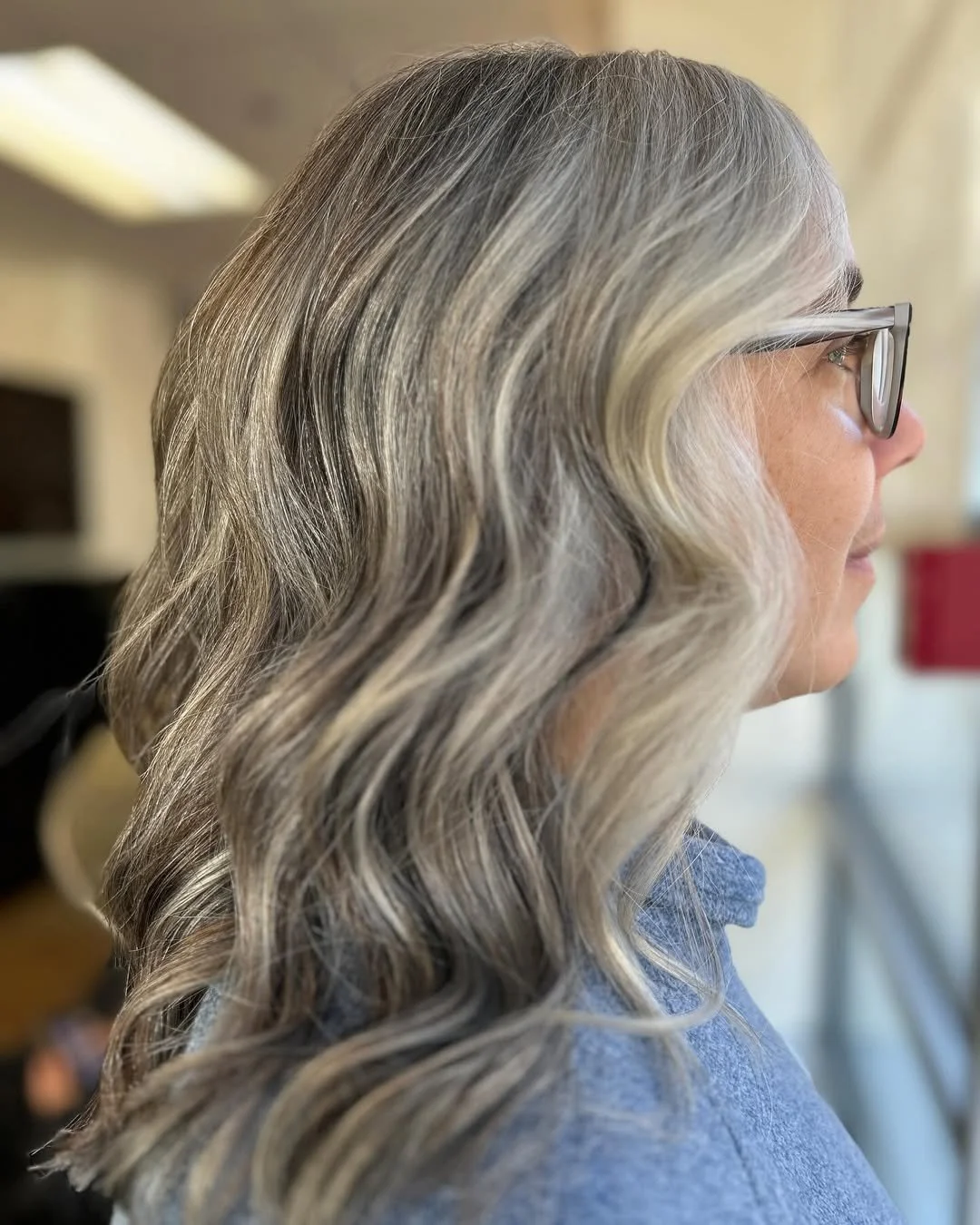 Profile side view of a woman with wavy, silver-gray hair, wearing glasses, indoors.