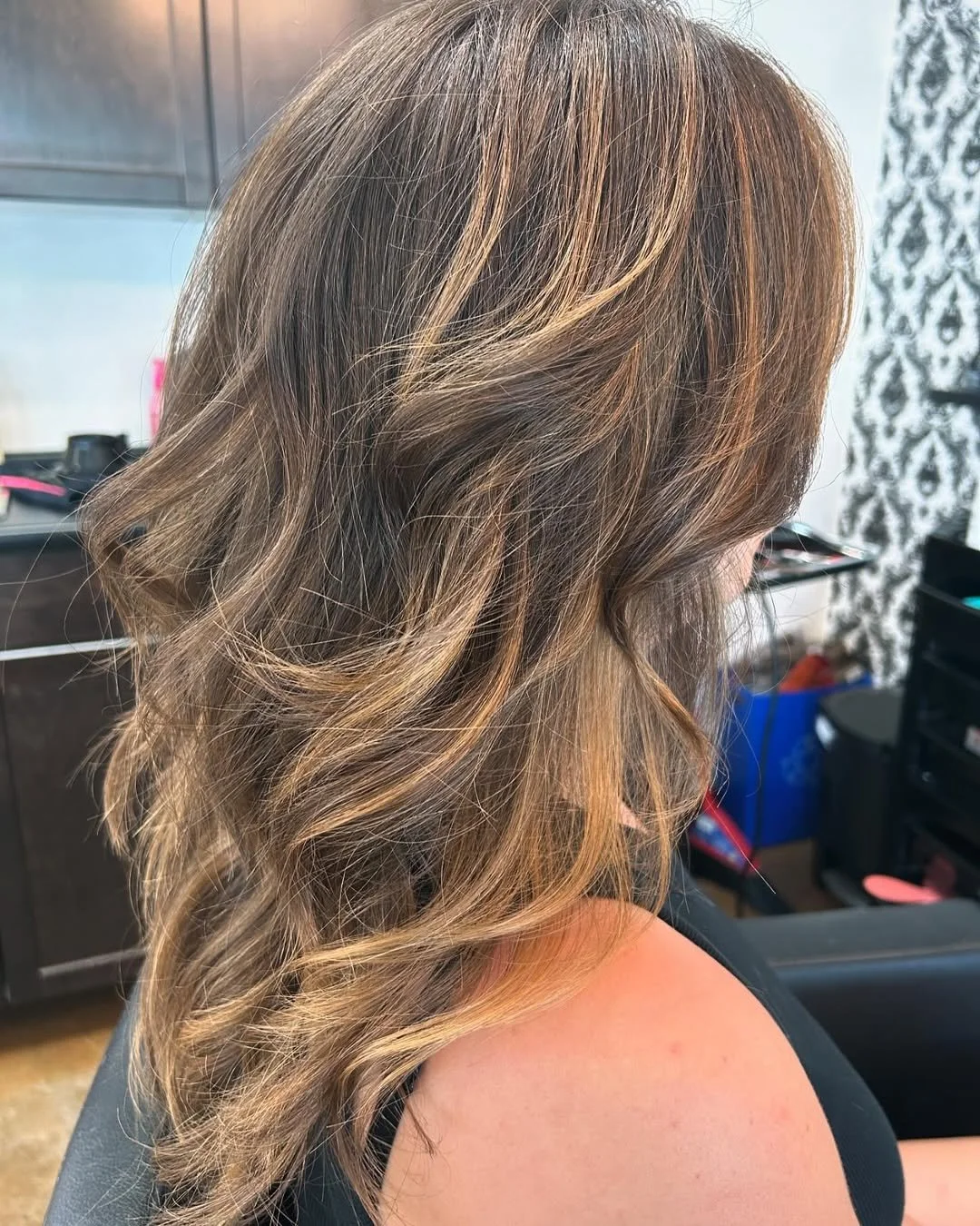 A woman with long, wavy hair styled in loose curls, featuring a blend of brown and blonde highlights, sitting in a salon chair.