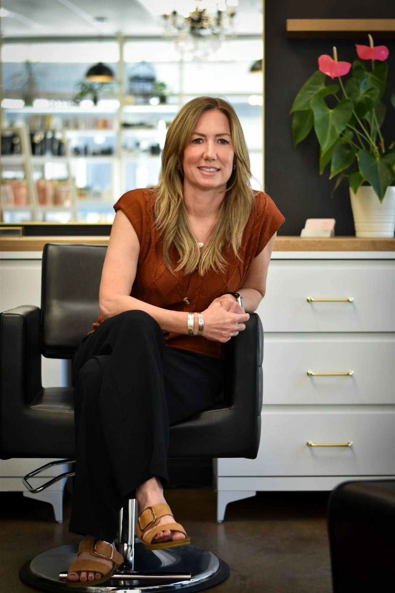 A woman with long, blonde hair sitting on a black office chair in a modern office or showroom. She is wearing a rust-colored sleeveless top, black pants, and tan sandals, with bracelets on her wrist. Behind her, there are shelves with various items and a large potted plant with pink flowers.