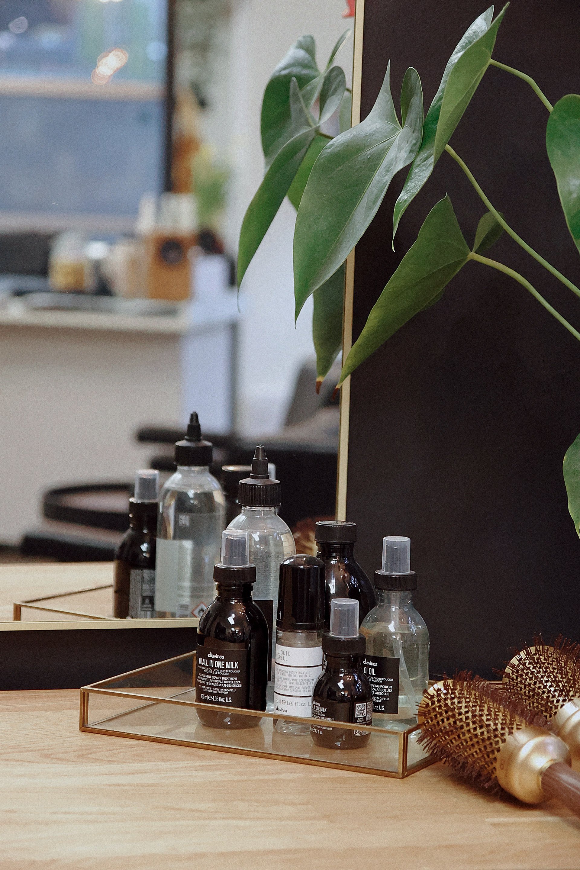 A wooden tray on a light wood table holds various skincare bottles and jars, some with droppers. Behind the tray, a mirror reflects a plant with broad green leaves. To the right, there is a hairbrush and a pearl hair accessory, with a blurred background of a room.