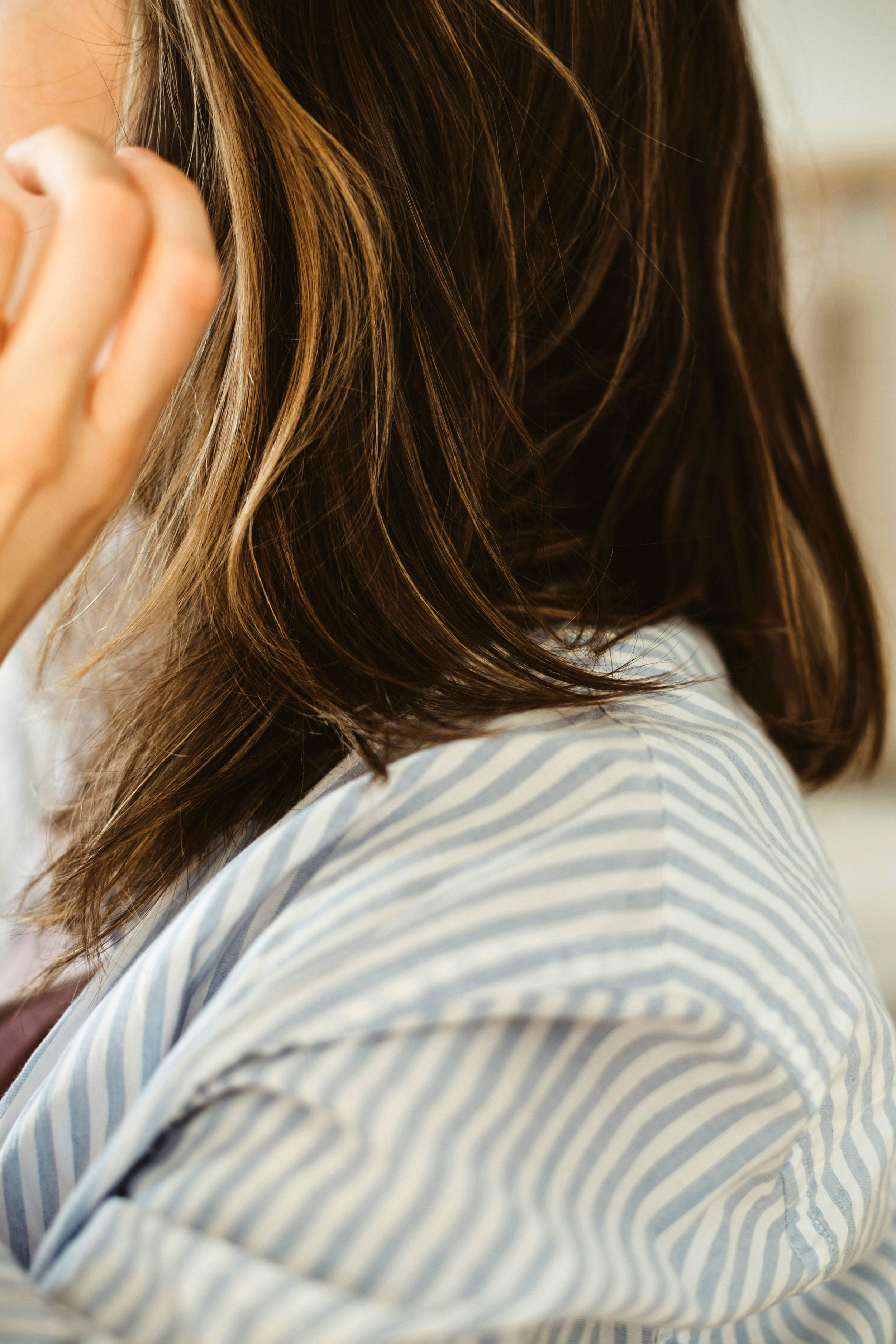 Close-up of a person with brown hair wearing a light-colored, striped shirt, touching their hair.