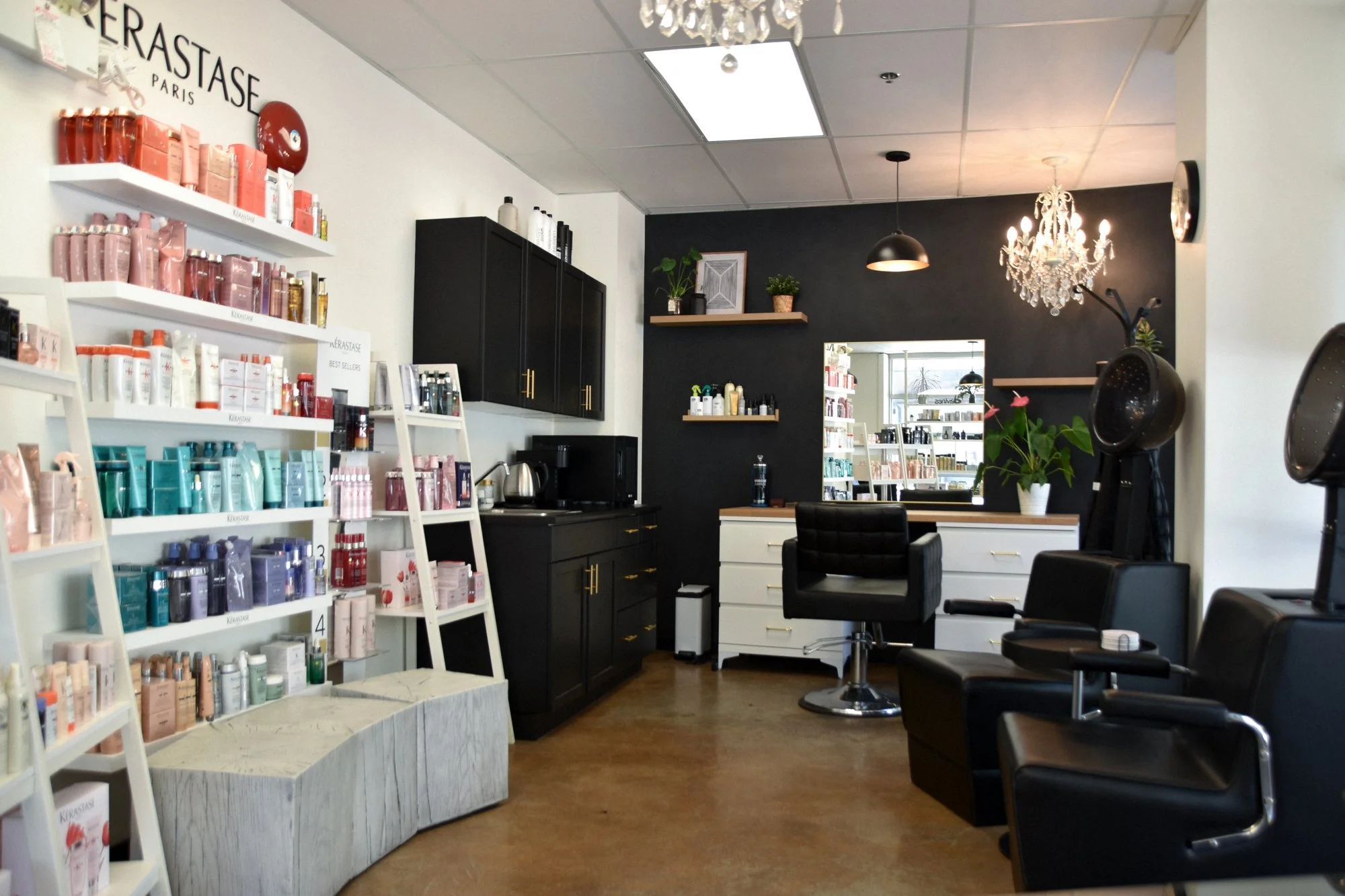 Interior of a hair salon or beauty studio with shelves of hair products, black cabinetry, a mirror, a chandelier, and hair styling chairs.