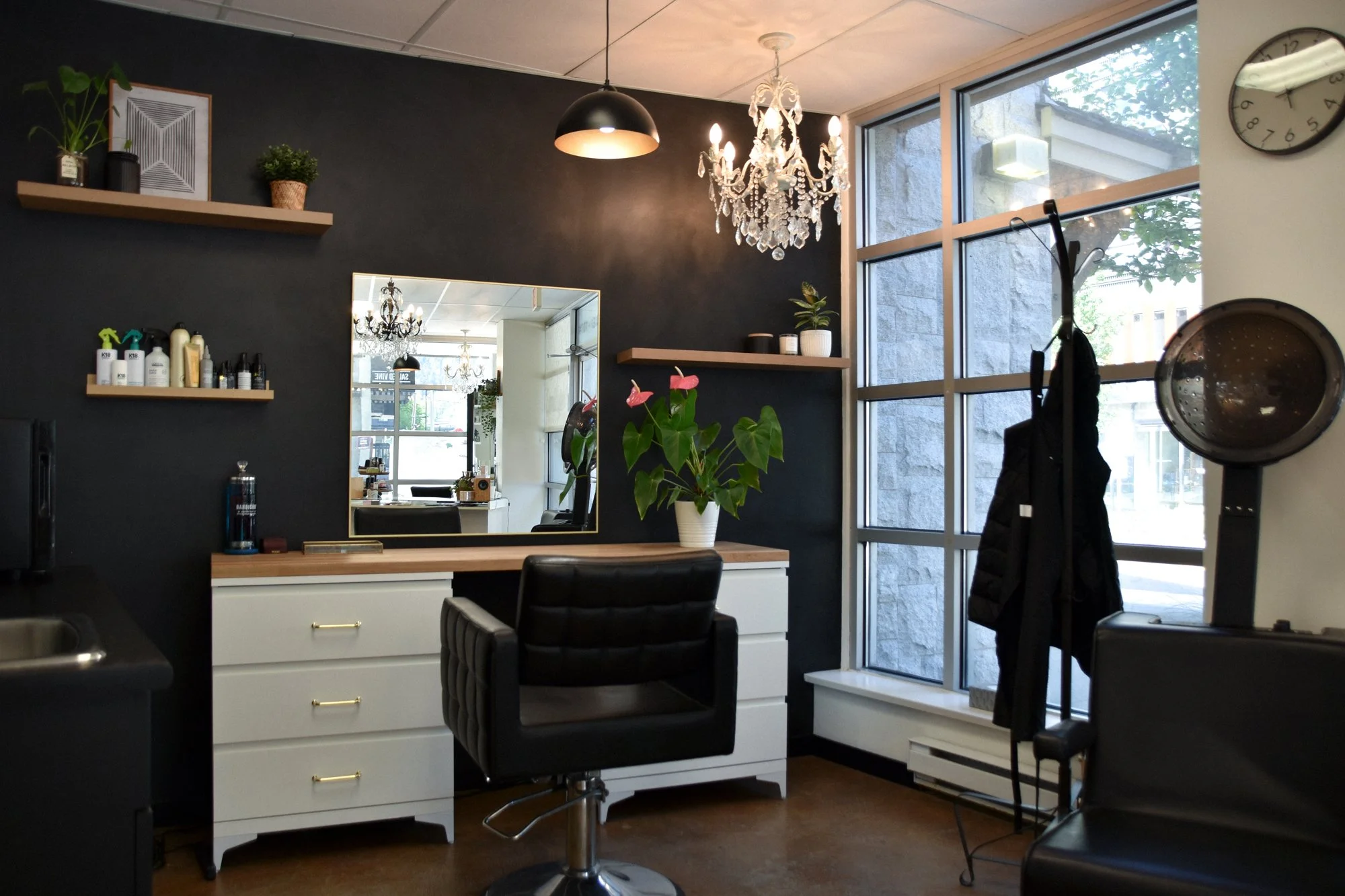 Interior of a modern hair salon with black walls, large windows, a chandelier, shelves with hair products, a mirror, and salon chairs.