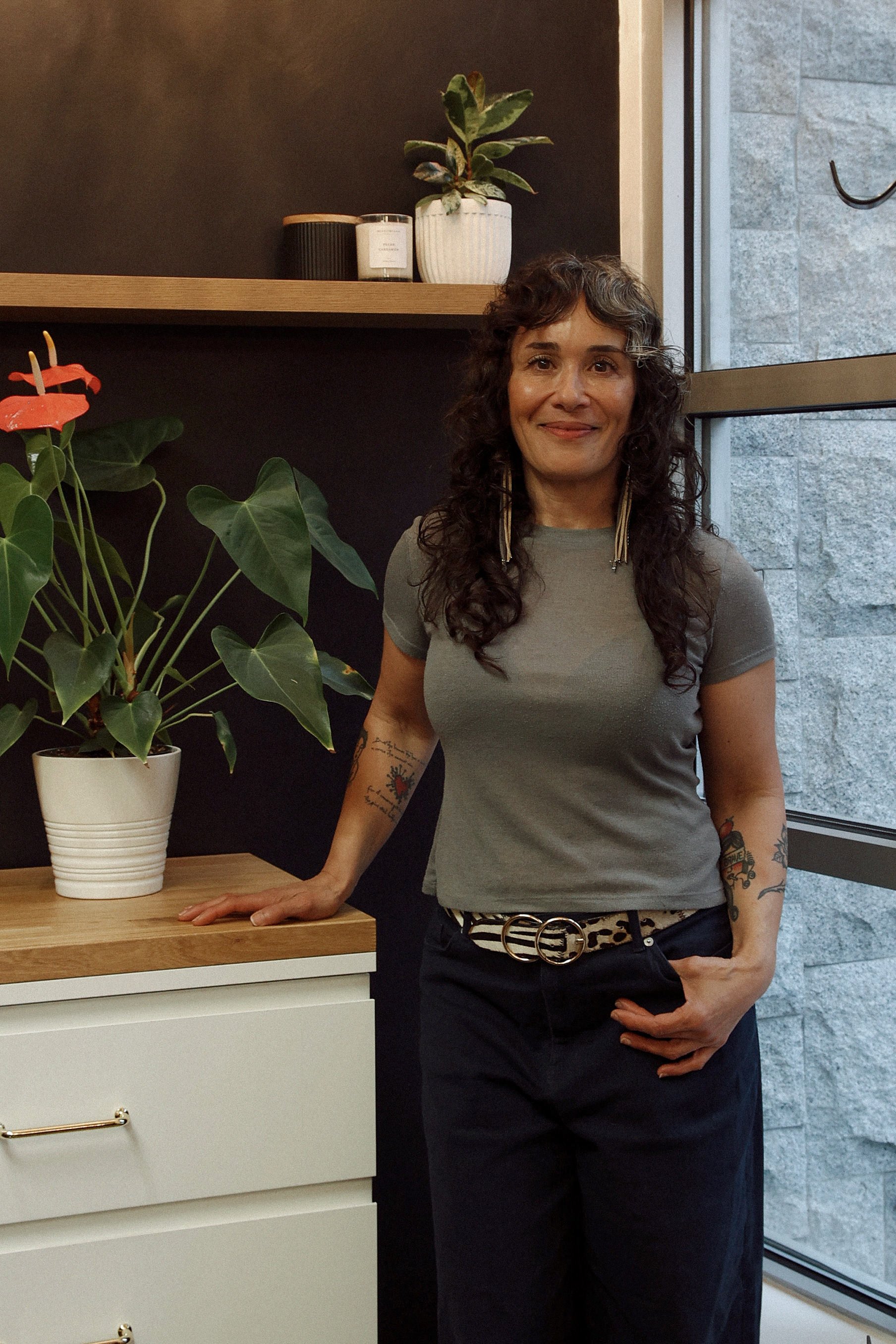 A woman with curly dark hair, tattoos on her arms, wearing a gray t-shirt and black pants with a leopard print belt, standing indoors near a wooden counter with a potted plant and a white drawer unit, next to a large window showing a gray brick wall outside.