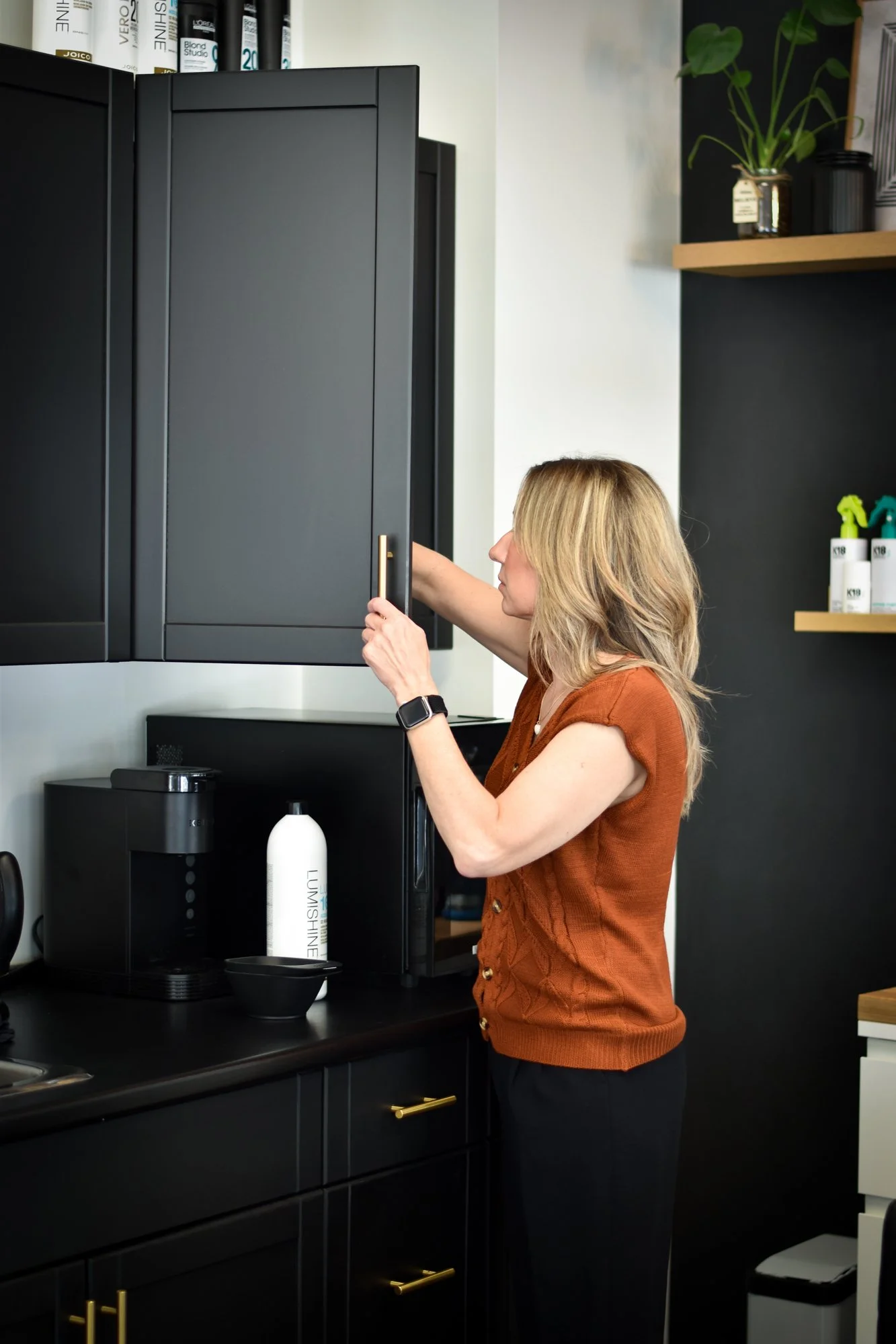 A woman with blonde hair wearing an orange sleeveless top preparing a drink in a modern kitchen with black cabinets, a coffee machine, and bottles on shelves.