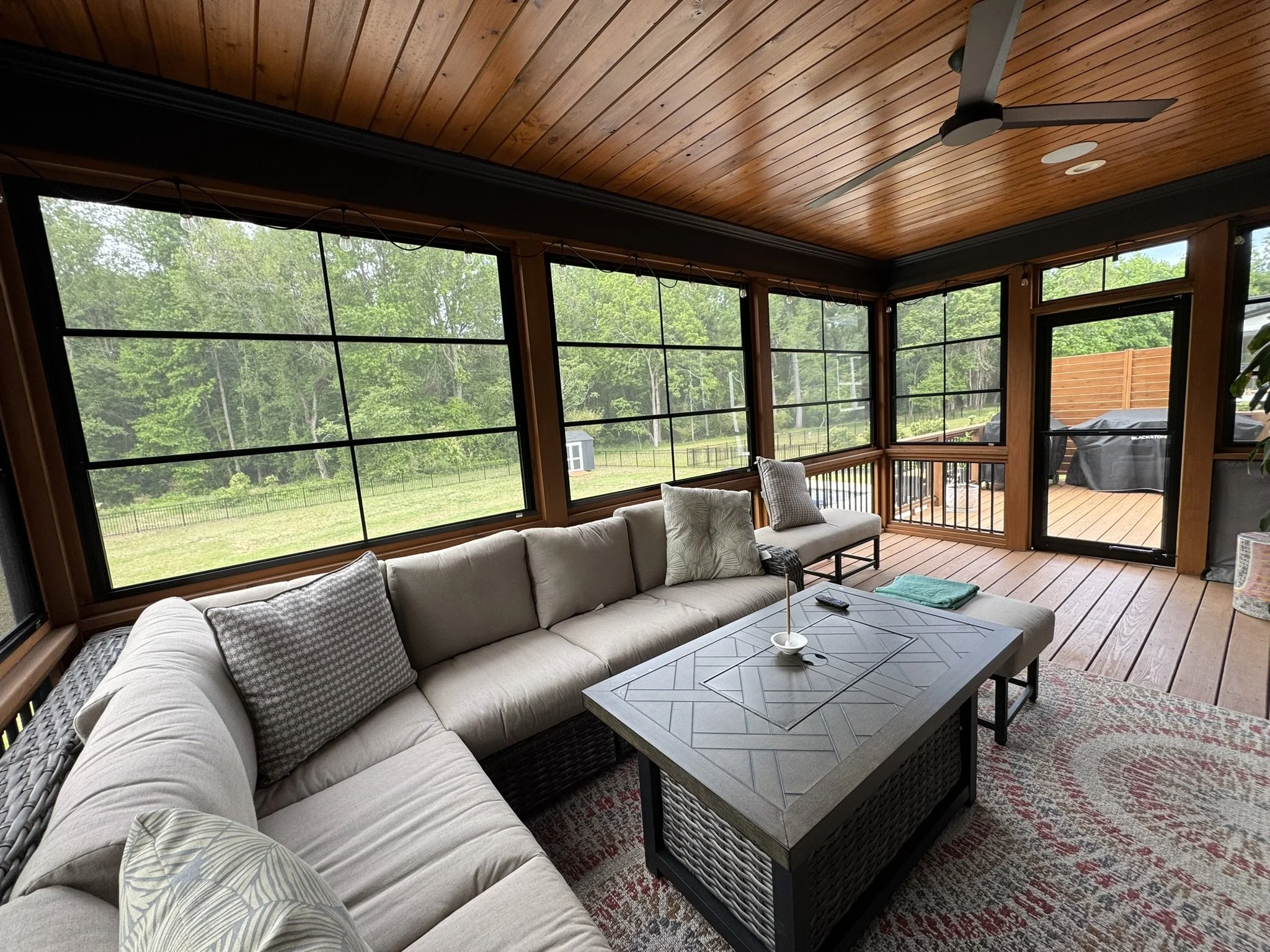 A screened porch with wooden ceiling and floor, furnished with a large beige sectional sofa with patterned and plain pillows, a square patio table with a geometric pattern, and a ceiling fan. The porch overlooks a green backyard with trees, a small s