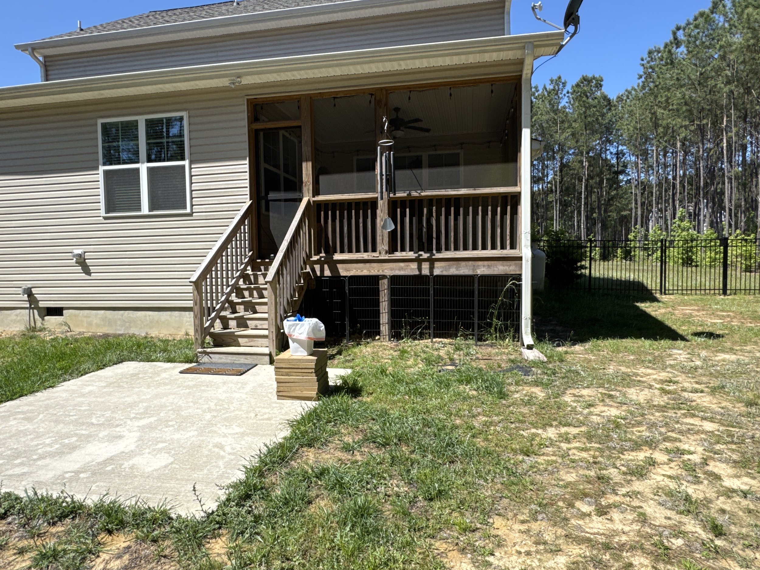 Rear view of a house with a screened porch, wooden stairs, and a small yard, surrounded by a black fence and trees under a clear blue sky.