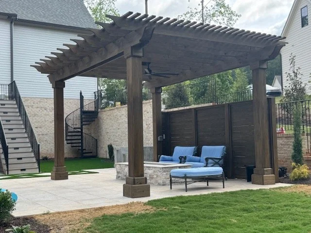 Backyard patio with a wooden pergola, outdoor seating with blue cushions, a concrete fire pit, and a brick retaining wall with stairs leading up to an upper level.